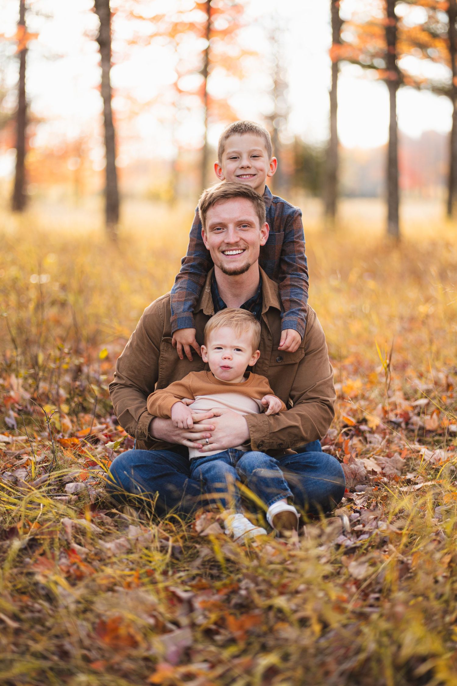 Smiling father sitting in autumn leaves holding two young sons in a golden fall forest setting.