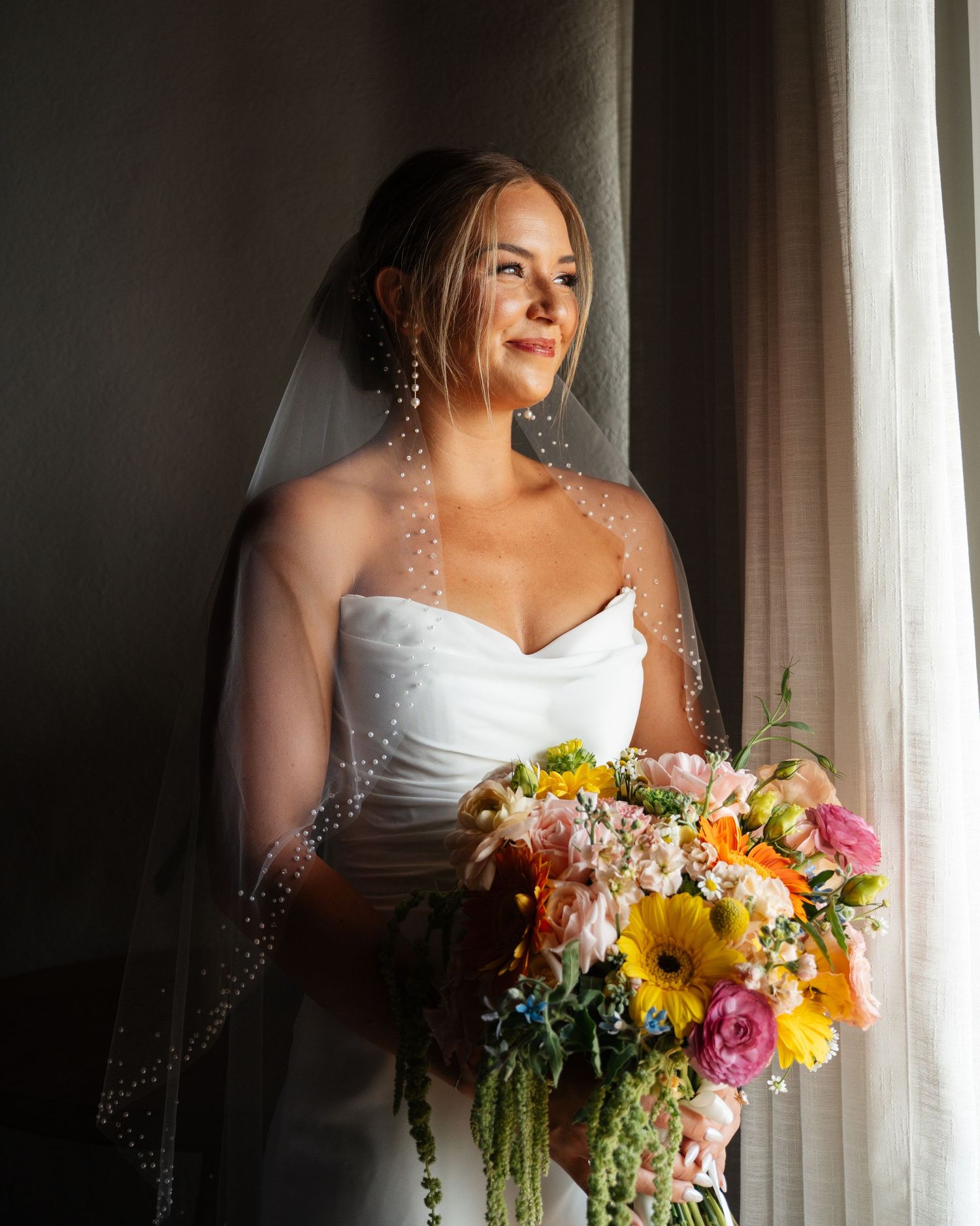 Elegant bride in white strapless gown holding colorful bouquet while gazing out a window.