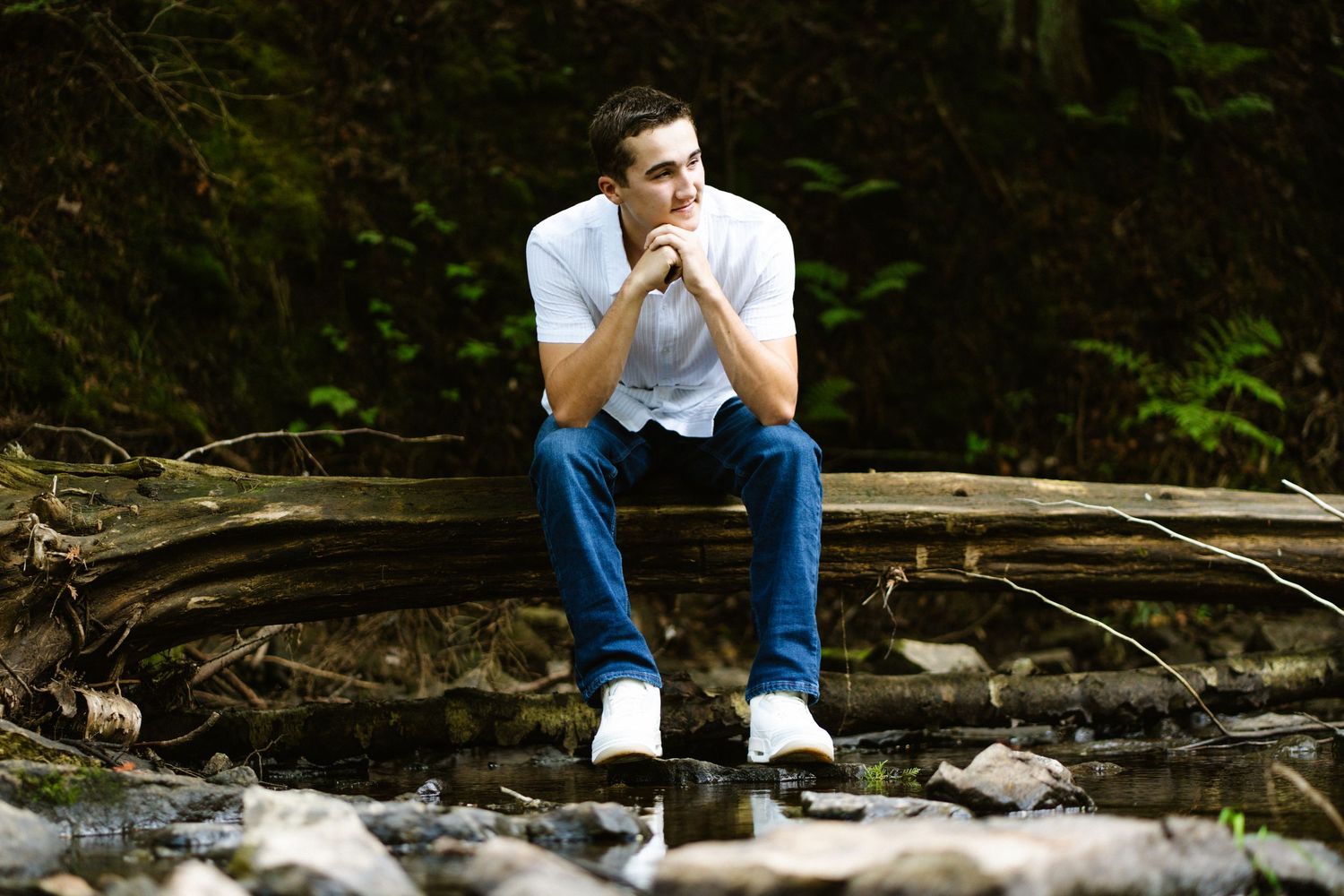 Young man in white shirt sitting thoughtfully on fallen log over rocky stream in forest.