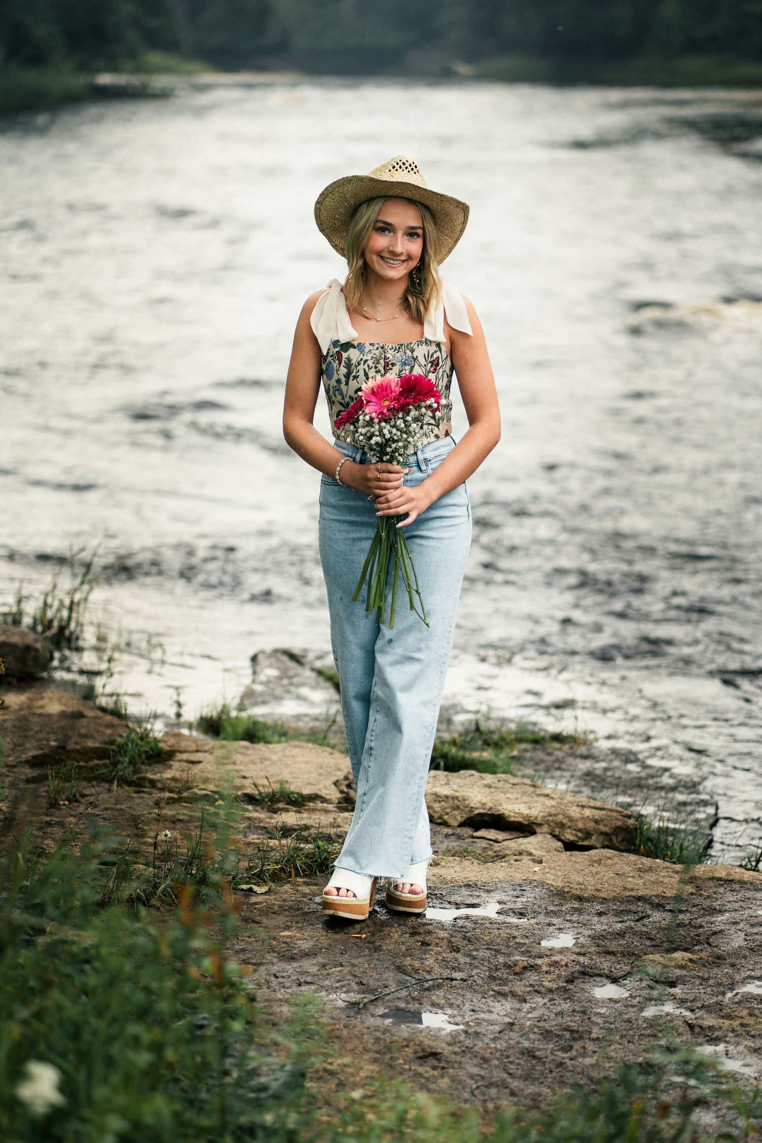 A smiling blonde woman in a straw hat and jeans holds a bouquet of flowers beside a flowing river.