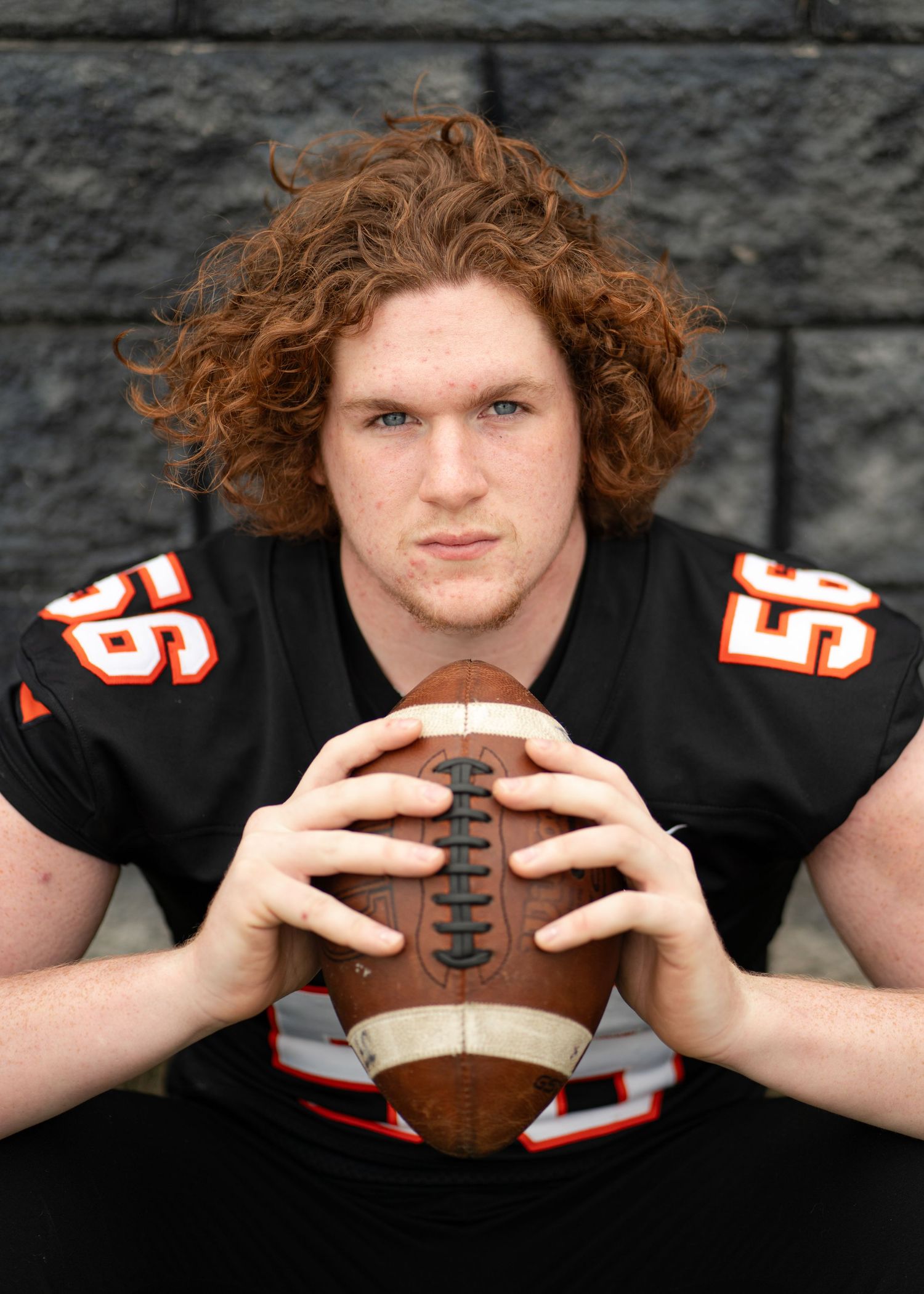 Curly red-haired football player in black jersey holding a football for a senior portrait.
