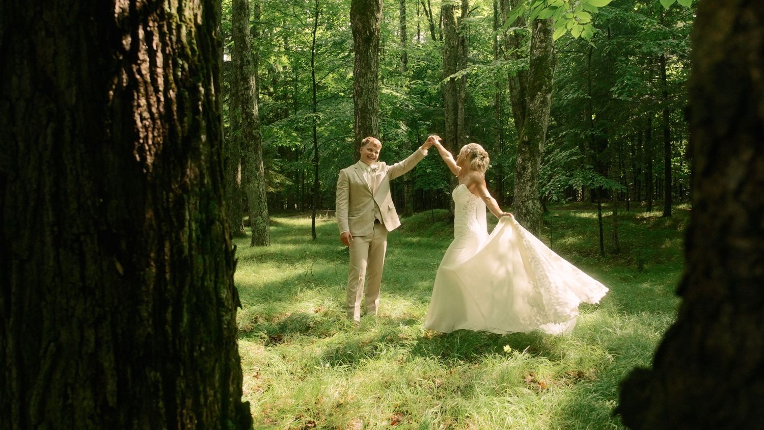 A groom spins his bride in a white gown between tall trees in a sunlit forest clearing.