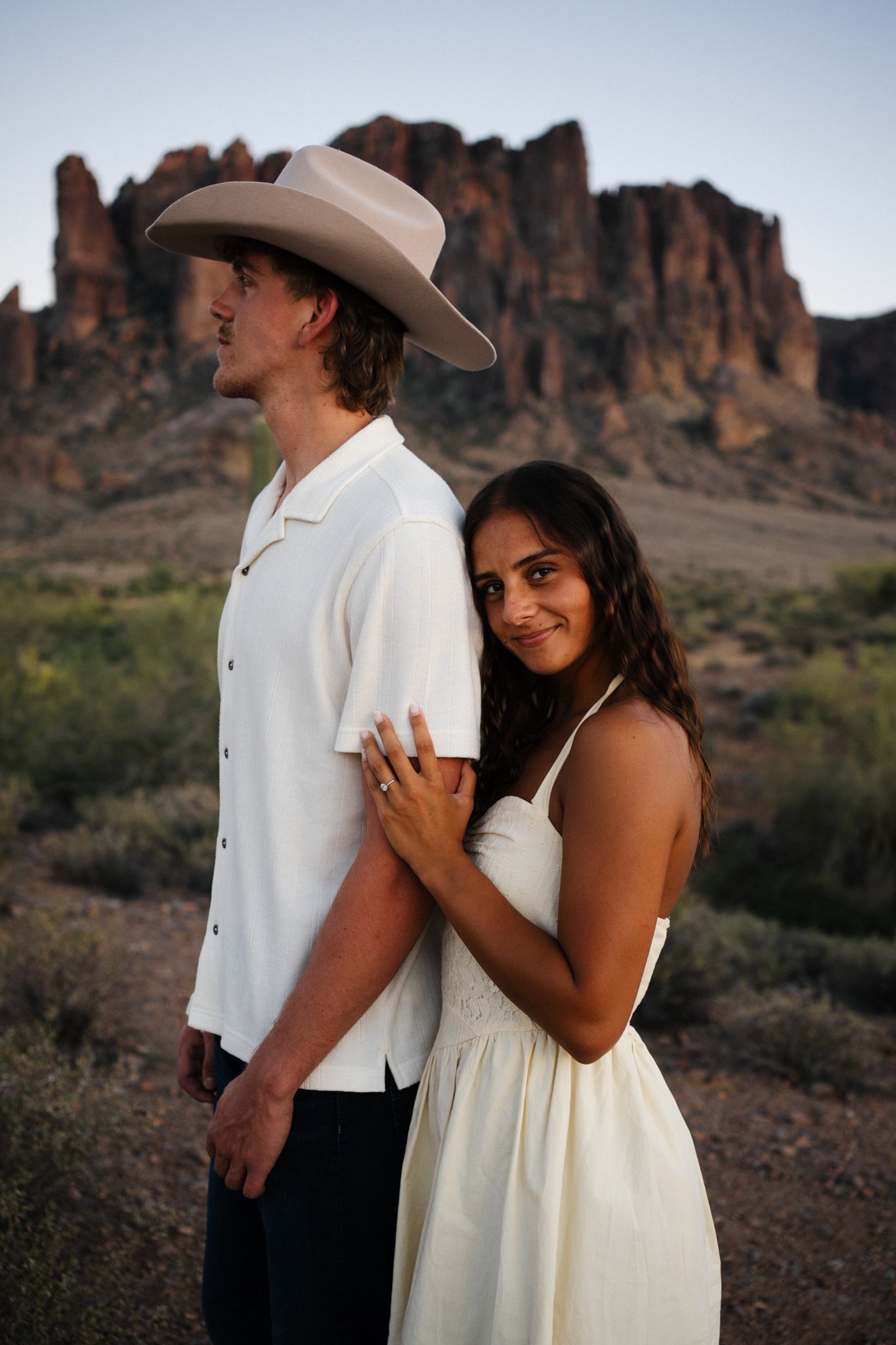 Woman in white dress embracing man in cowboy hat with rocky desert mountains behind them.