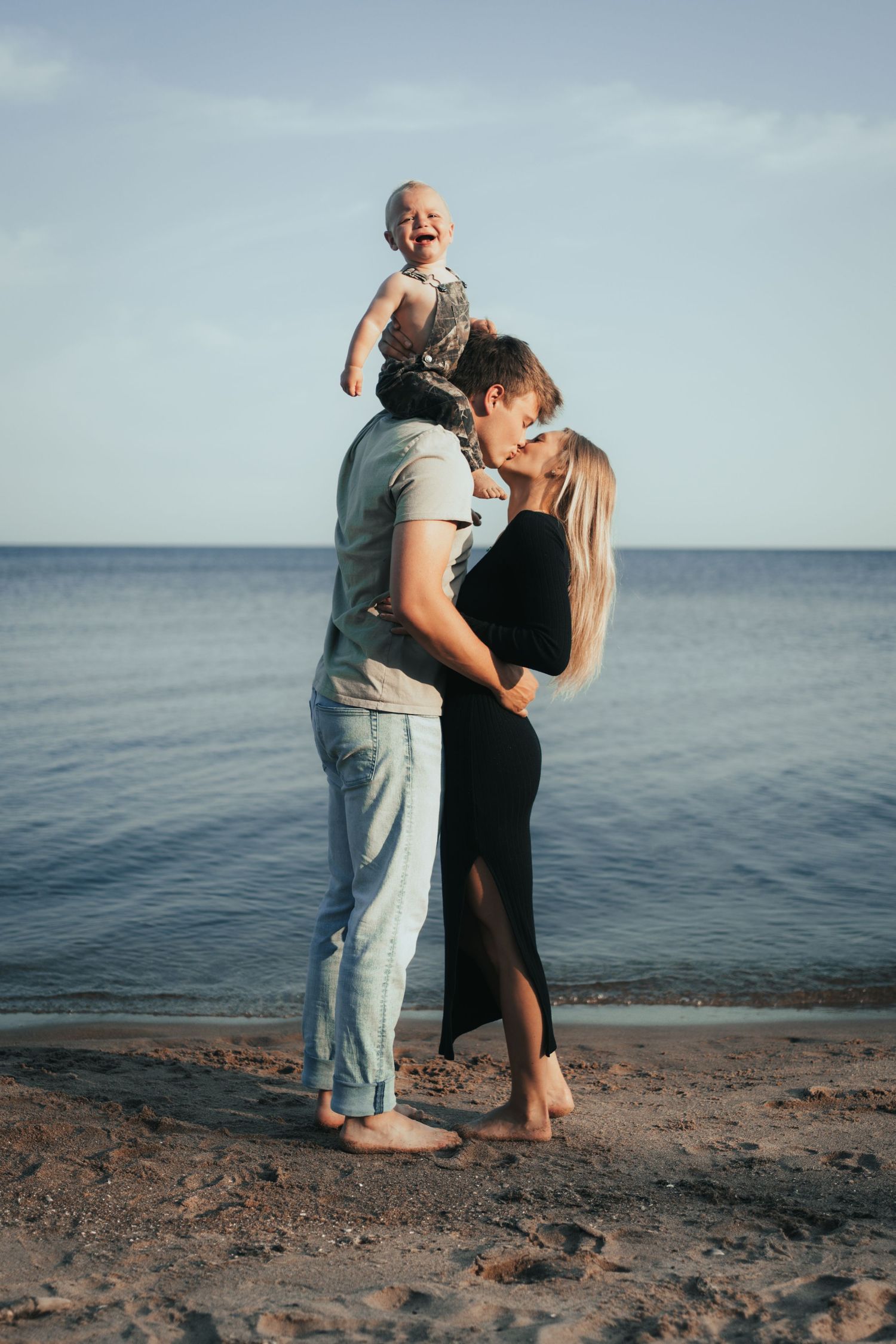 Joyful family at the beach with father holding laughing baby on shoulders while mother smiles nearby.