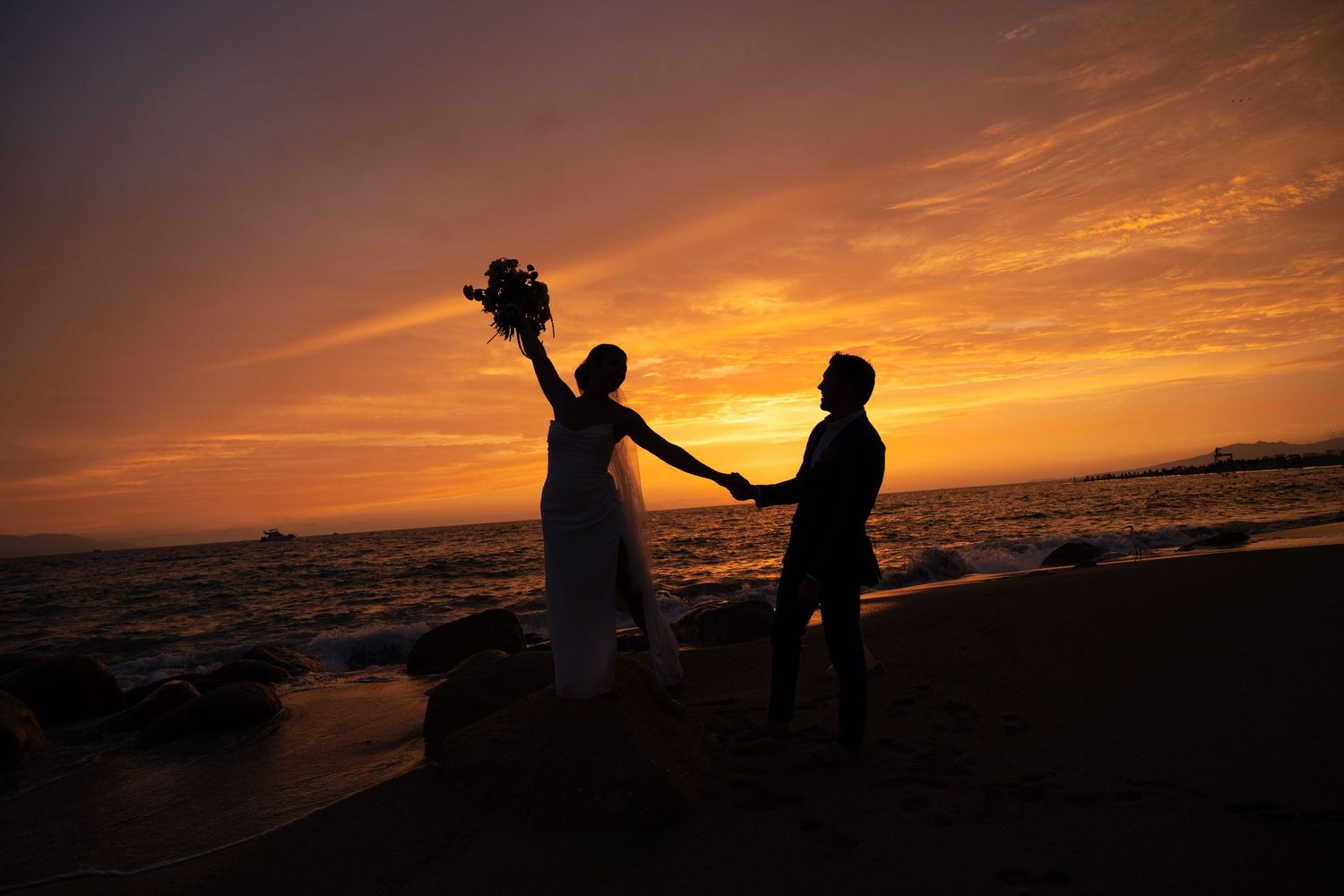 Silhouette of bride and groom holding hands on beach at stunning golden sunset with bouquet raised.