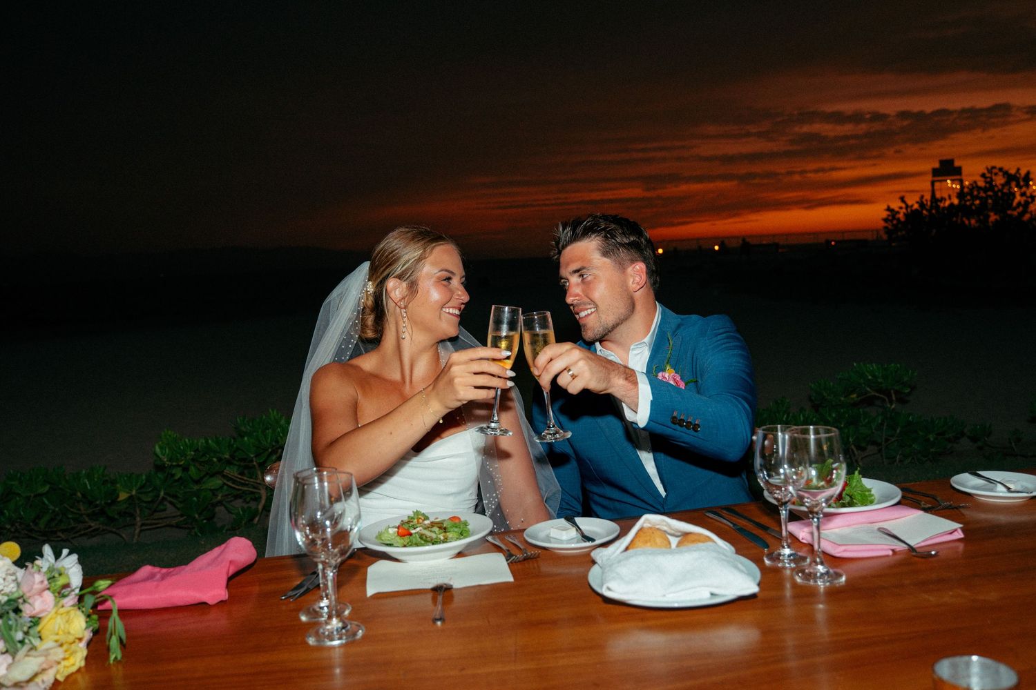 Newlyweds toast champagne at intimate beachside dinner table under a vibrant red and orange sunset sky.