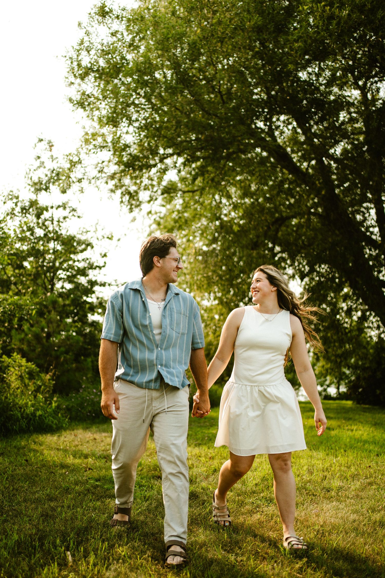 Happy couple holding hands and laughing while walking through lush green park at sunset.