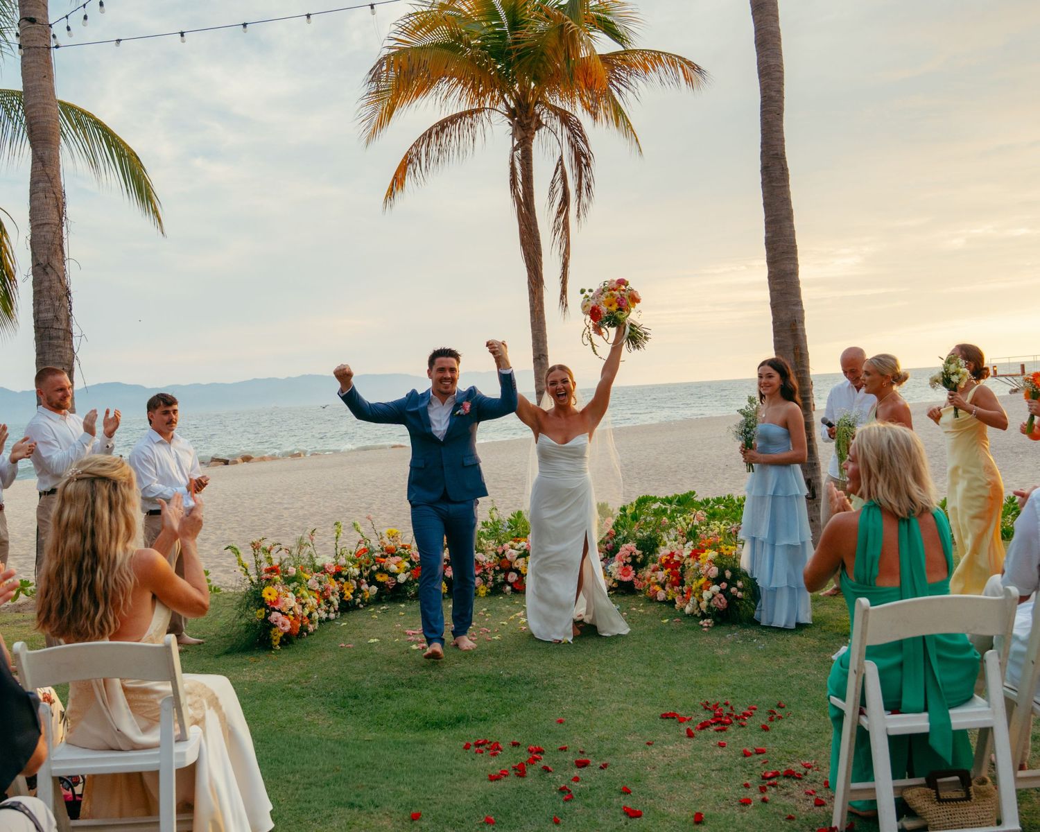 Joyful newlyweds raise their arms as they walk back down the aisle at a beachside sunset ceremony.