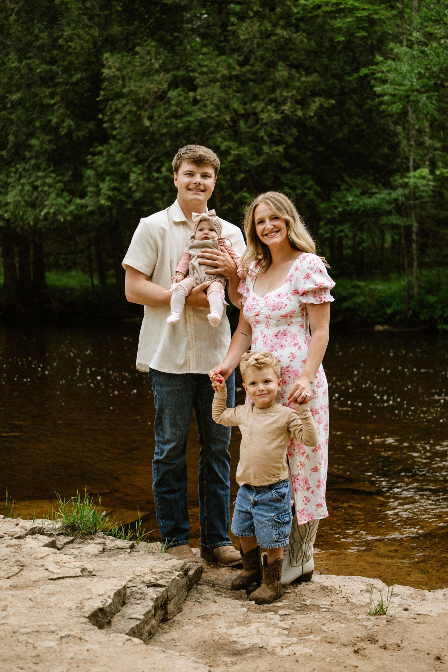 Young family of four poses by a serene forest river, parents holding toddler and newborn baby outdoors.