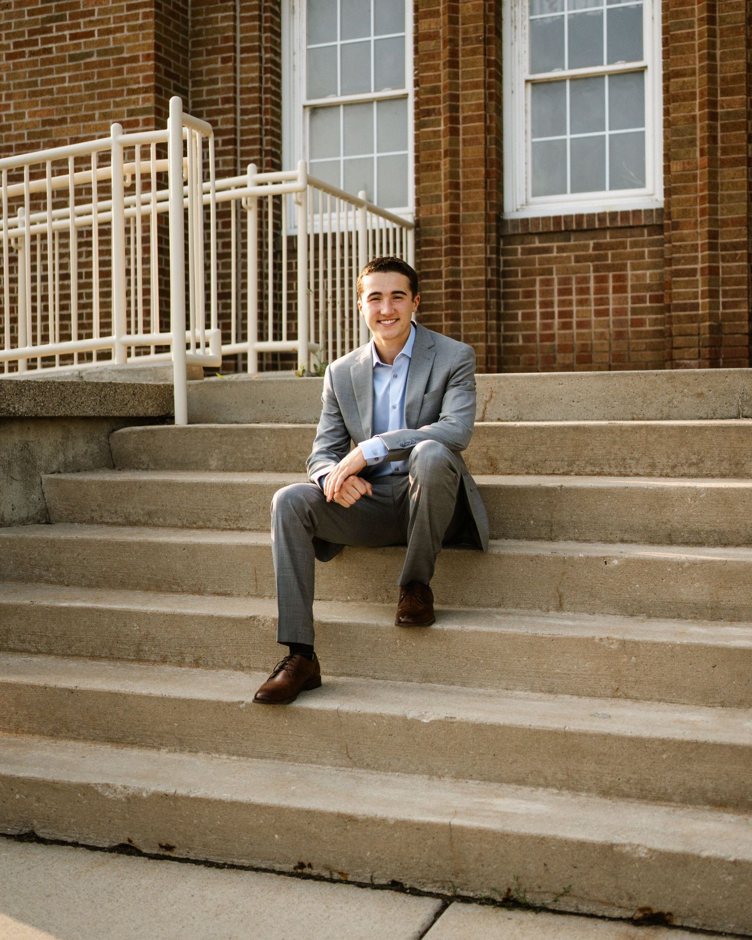 Smiling young man in gray suit sitting on brick building steps for senior portrait photo.