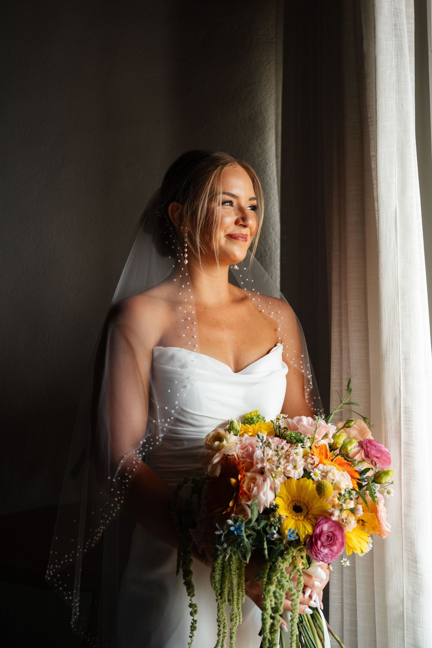 Radiant bride in white gown and veil holds colorful bouquet while gazing out sunlit window.