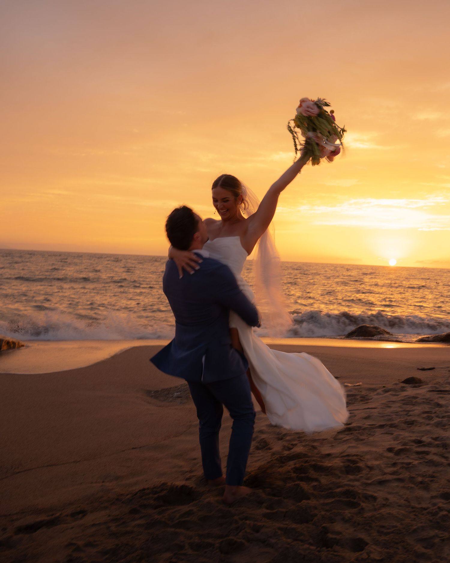 Groom lifts bride holding bouquet high on sandy beach during golden sunset wedding photo session by the ocean.