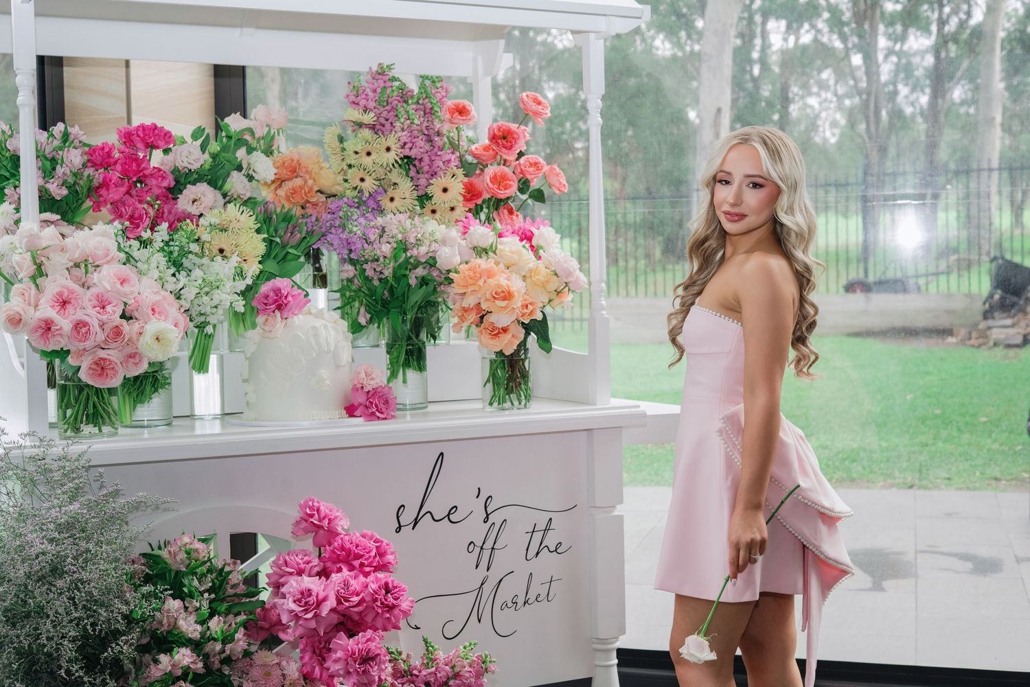A floral display stand decorated with vibrant pink and peach flowers sits on a porch at a special event.