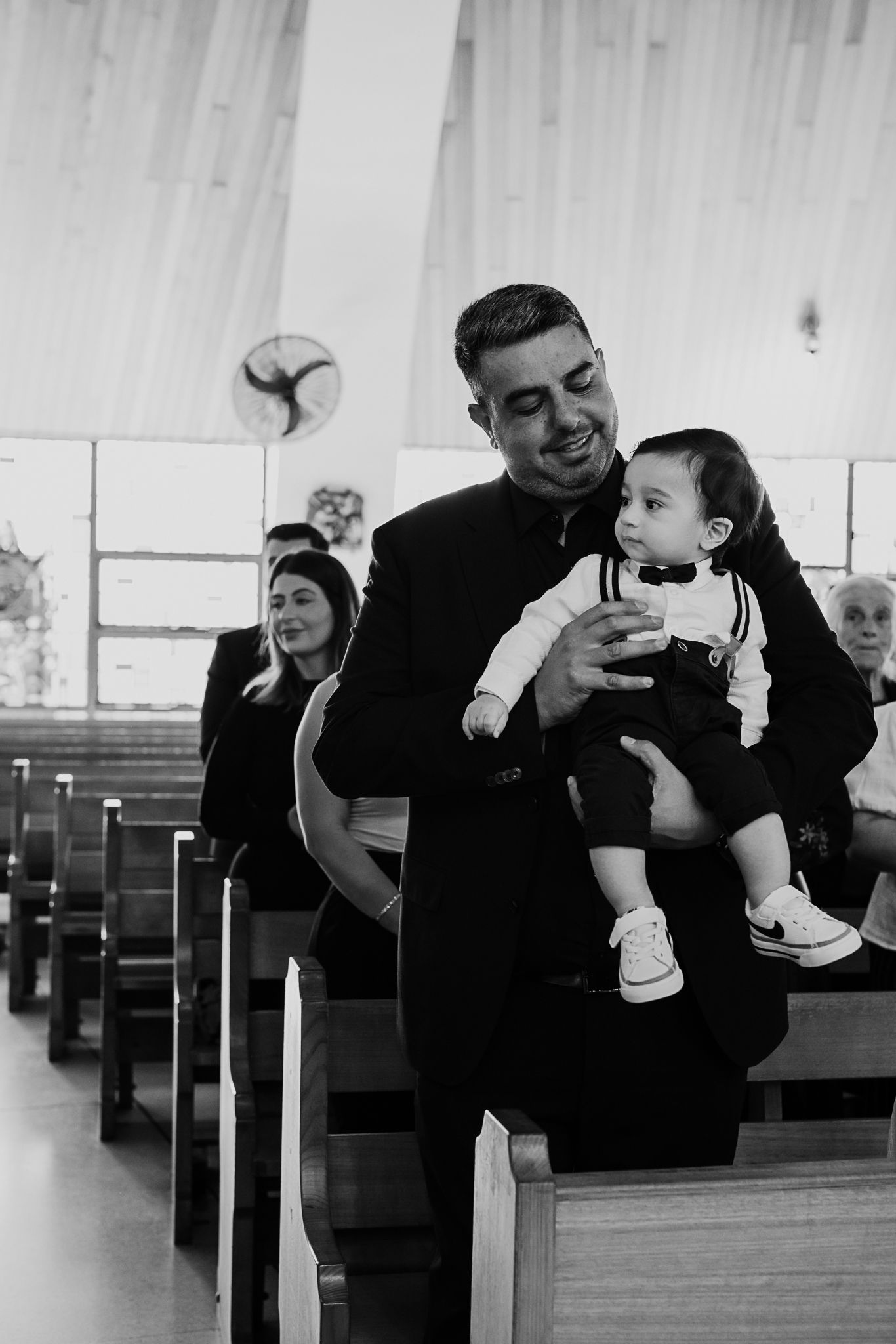 Black and white photo of priest holding baby in church pews.
