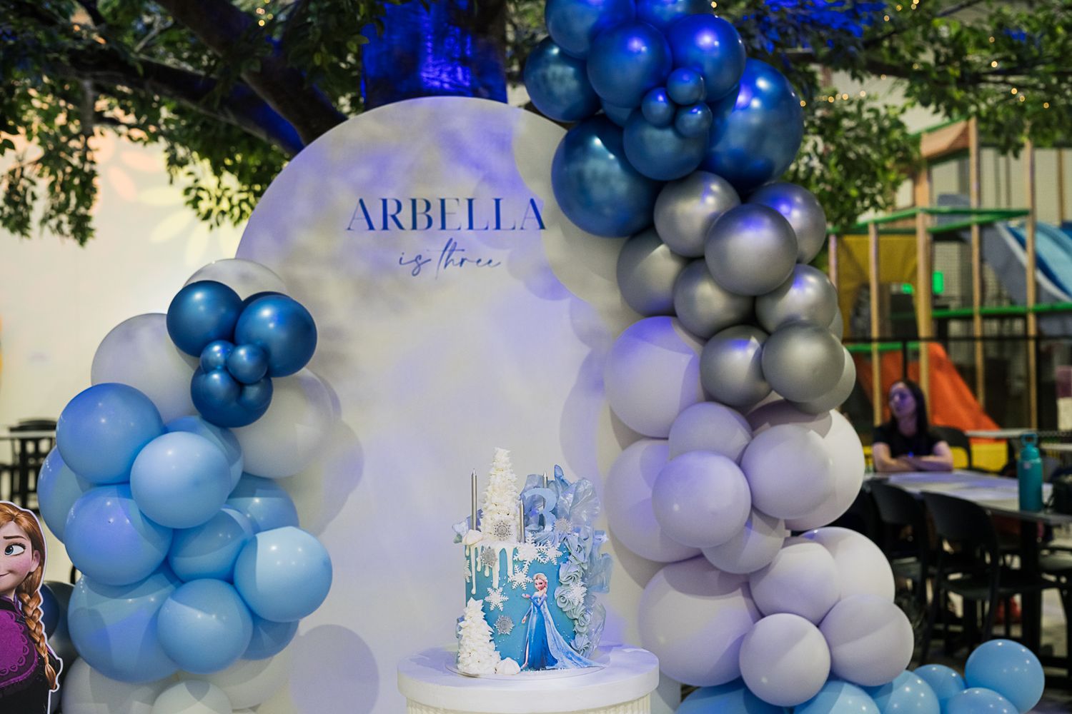 Frozen-themed party decoration with blue and silver balloon arch and white backdrop.