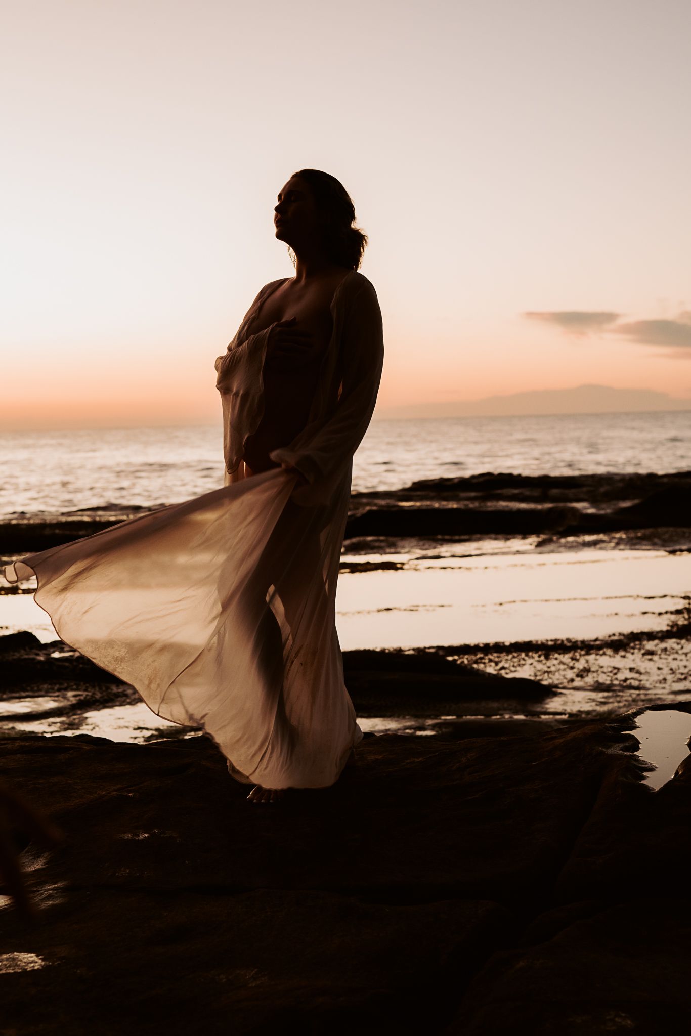 Silhouette of a figure in flowing dress against sunset ocean backdrop on rocky shore.