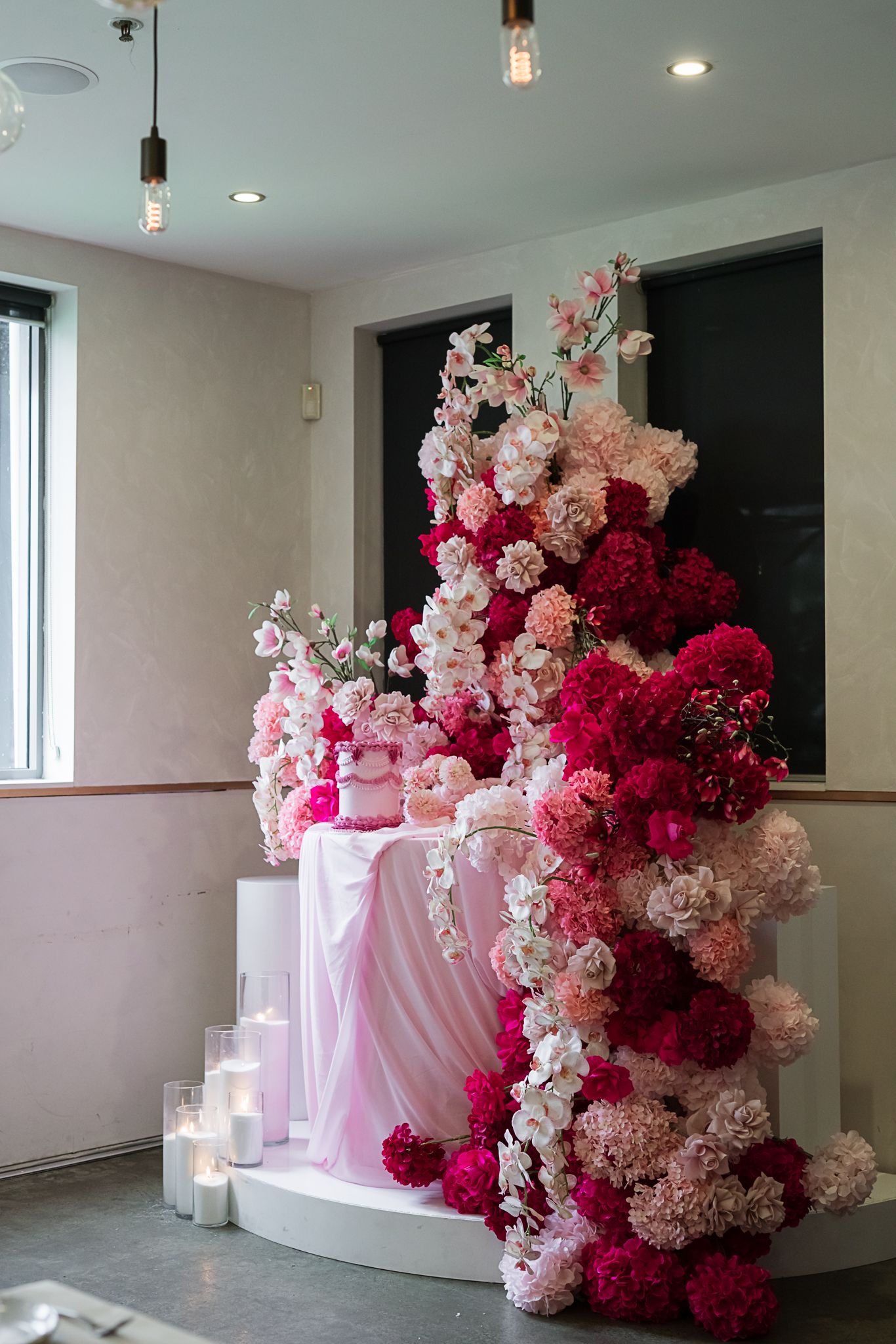 Elegant wedding display featuring a cascading floral arrangement in pink and burgundy with white candles on draped fabric.
