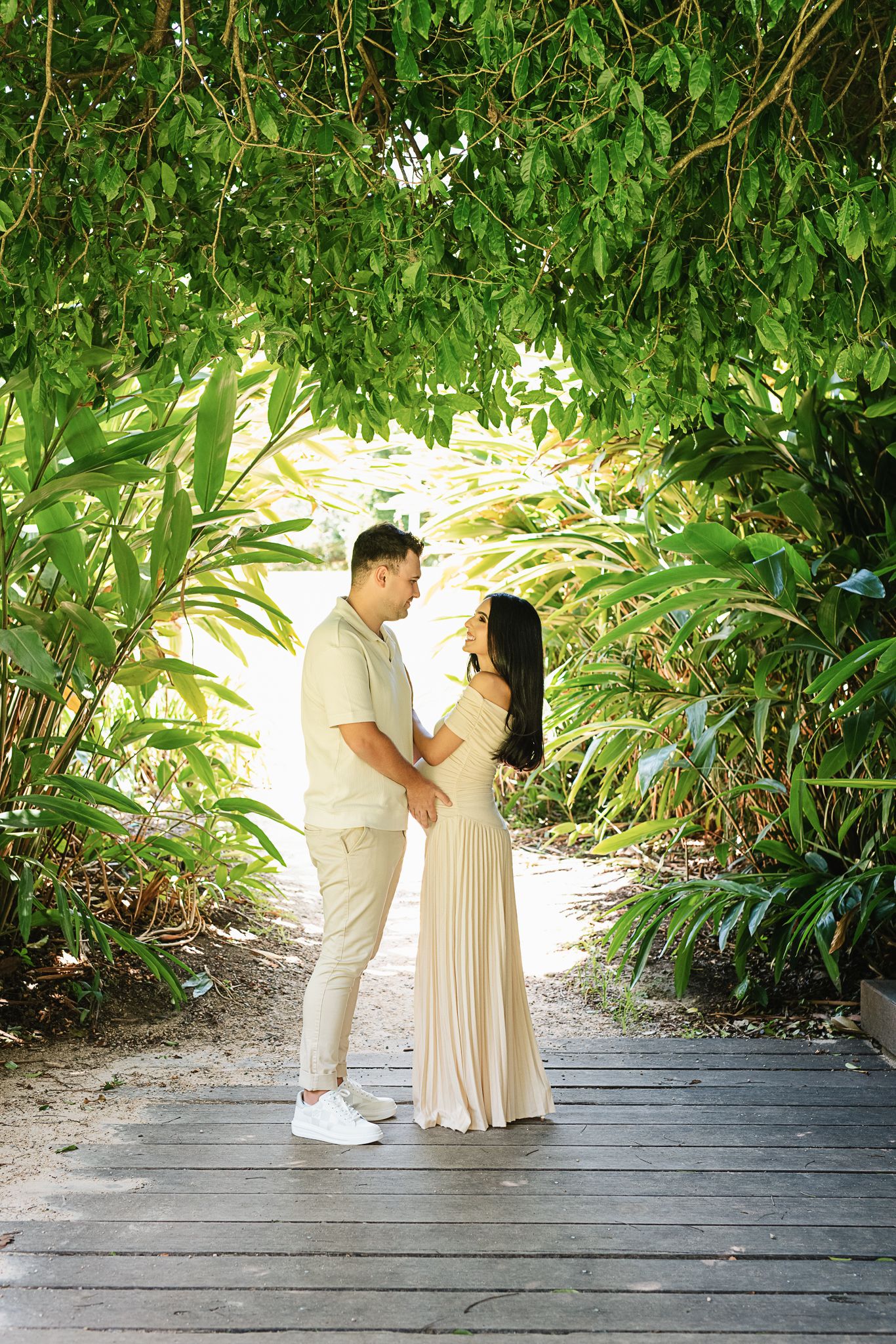 Expecting parents pose on a wooden walkway surrounded by tropical greenery.