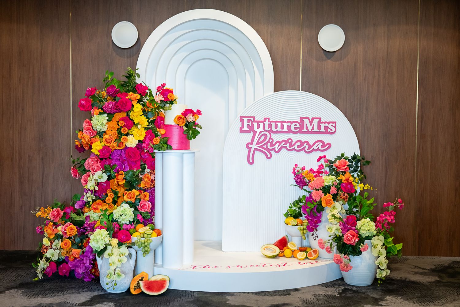 White arched backdrop with vibrant floral arrangements and pink text for a bridal event.