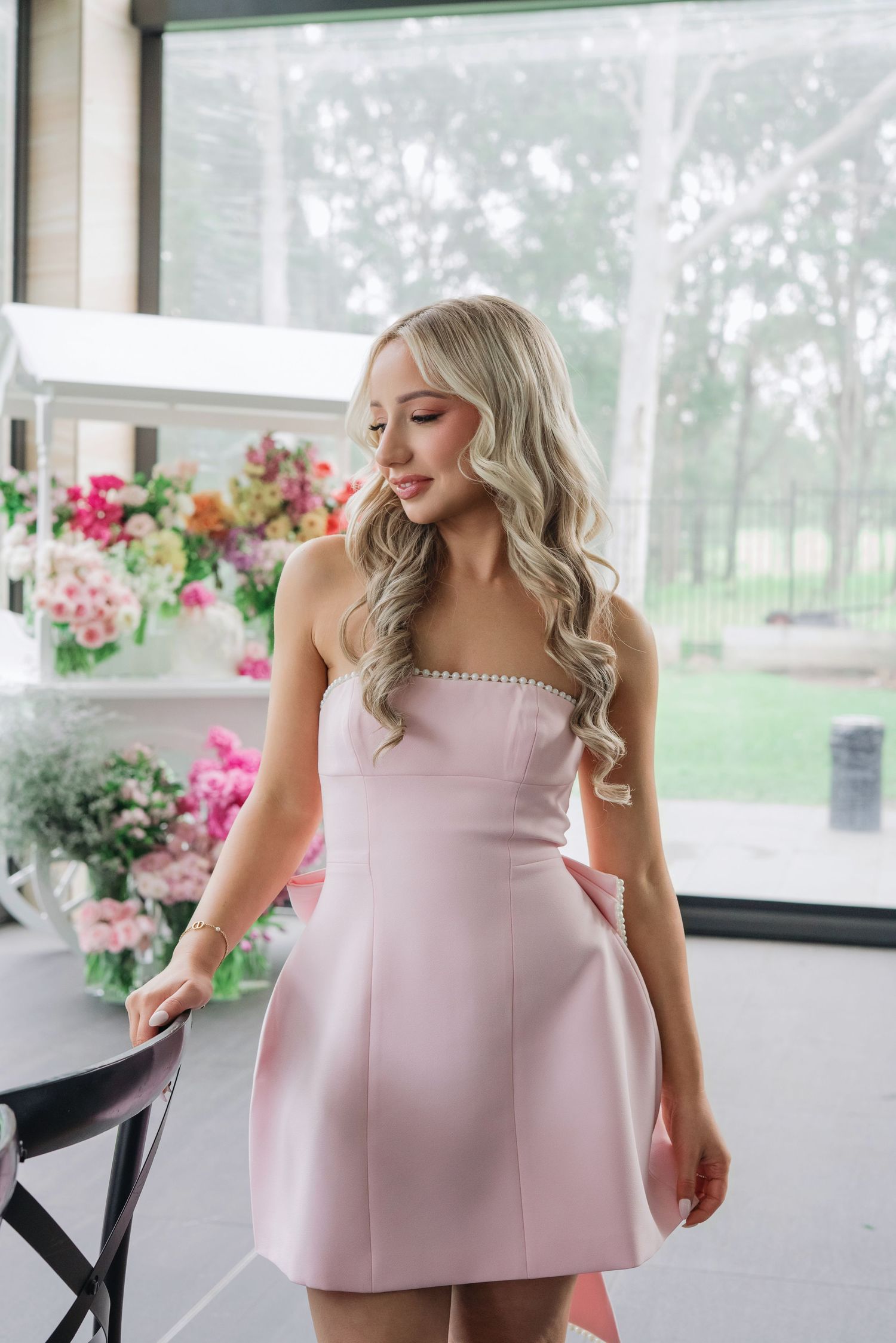 A woman in a strapless light pink cocktail dress poses on a porch with flowers in the background.
