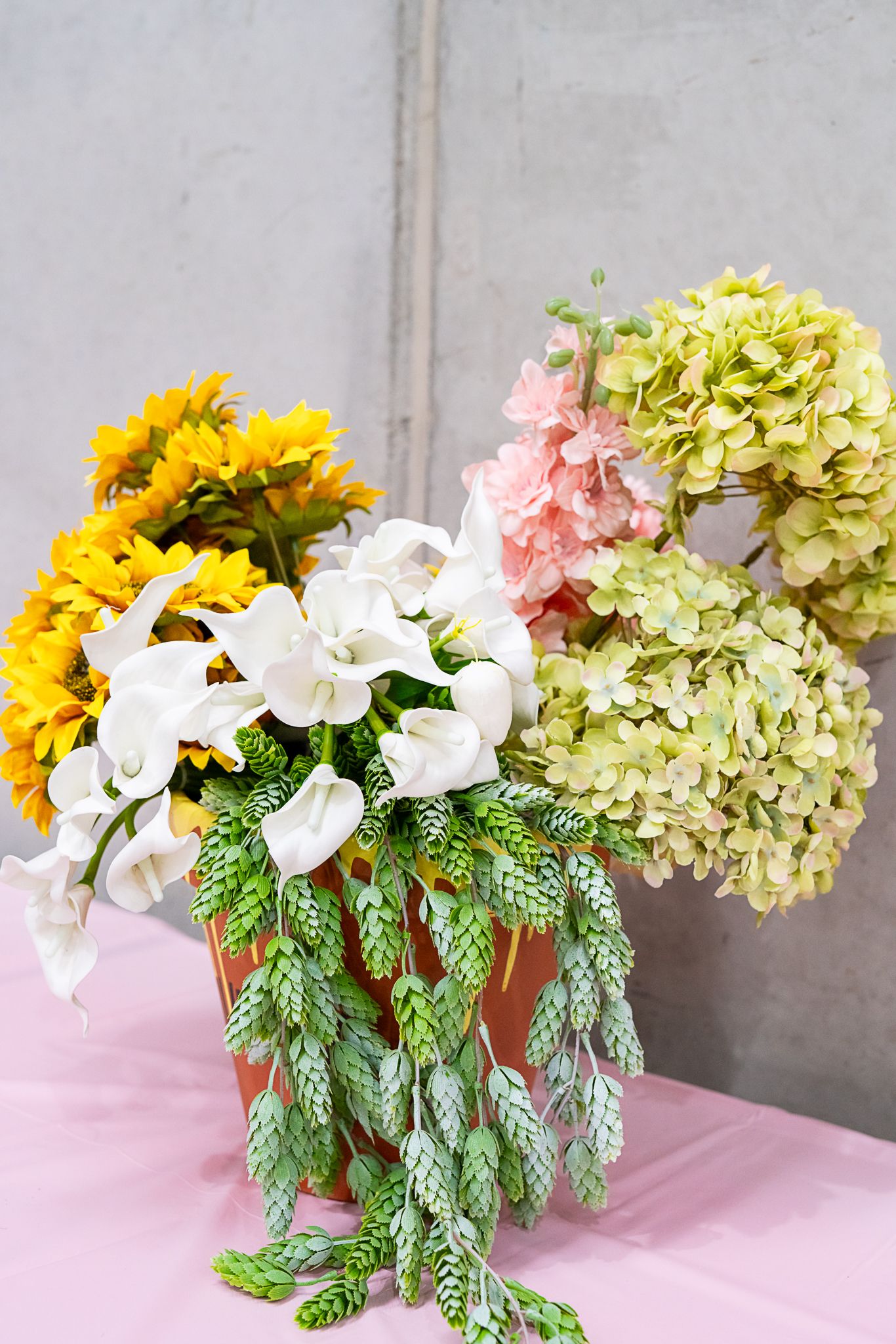 Elegant floral arrangement with sunflowers, calla lilies, and hydrangeas on pink tablecloth.