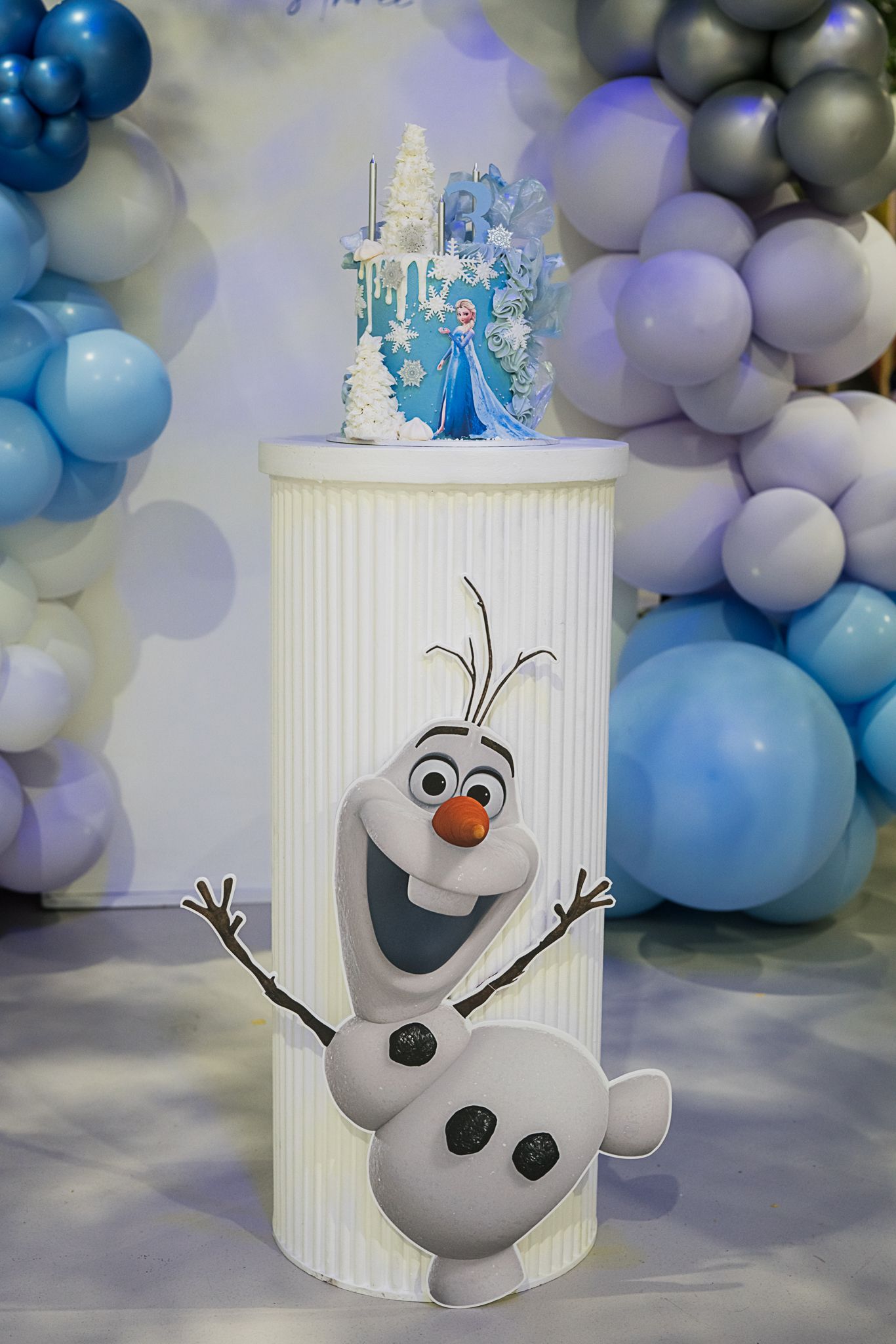 Olaf-themed party decoration with cake stand and blue balloon arch backdrop.