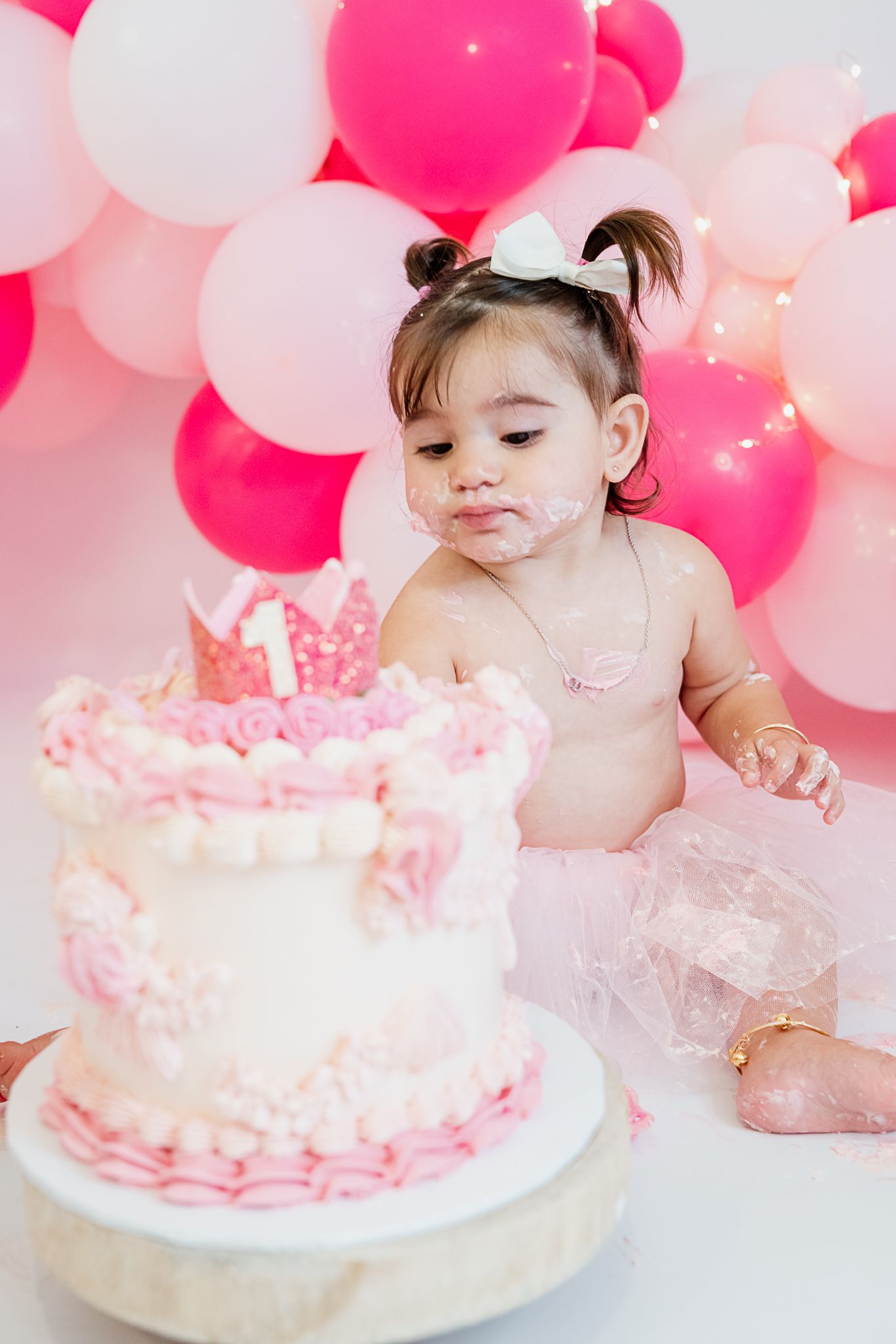 Baby in a pink tutu celebrating first birthday with cake and pink balloon backdrop.