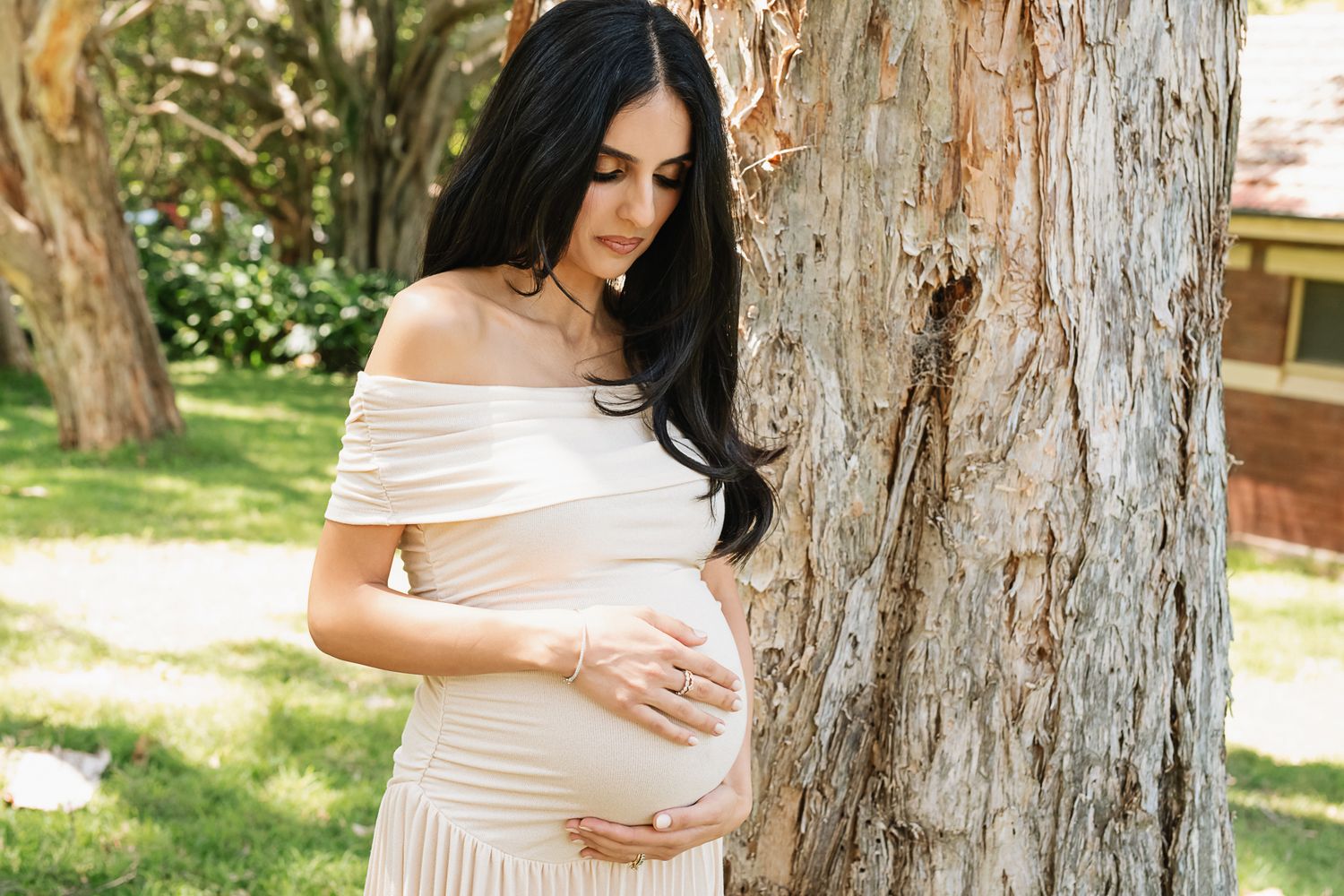 Side view of pregnant woman in cream dress standing by textured tree bark in sunlit garden.