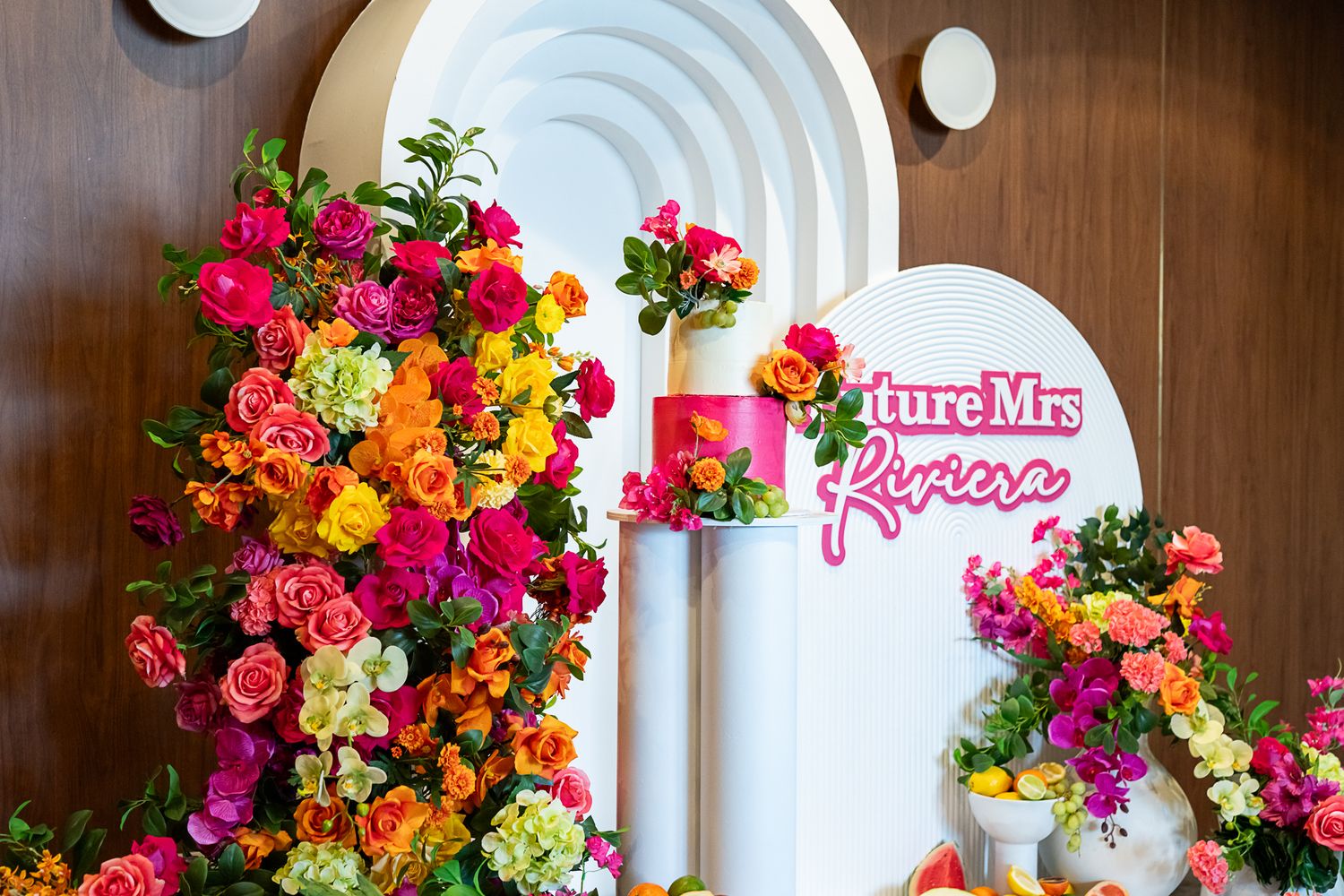 White archway decorated with vibrant pink, orange and yellow flowers and a pink neon sign for a wedding event.