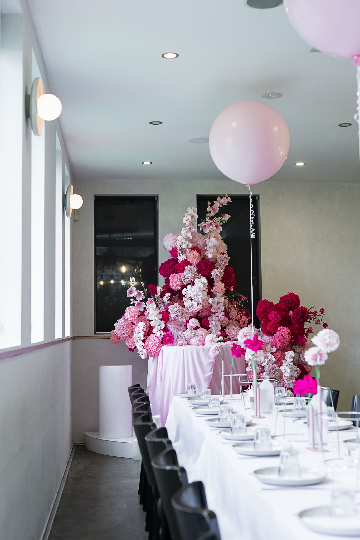 Long white dining table with pink balloon and floral arrangements in an elegant event space.