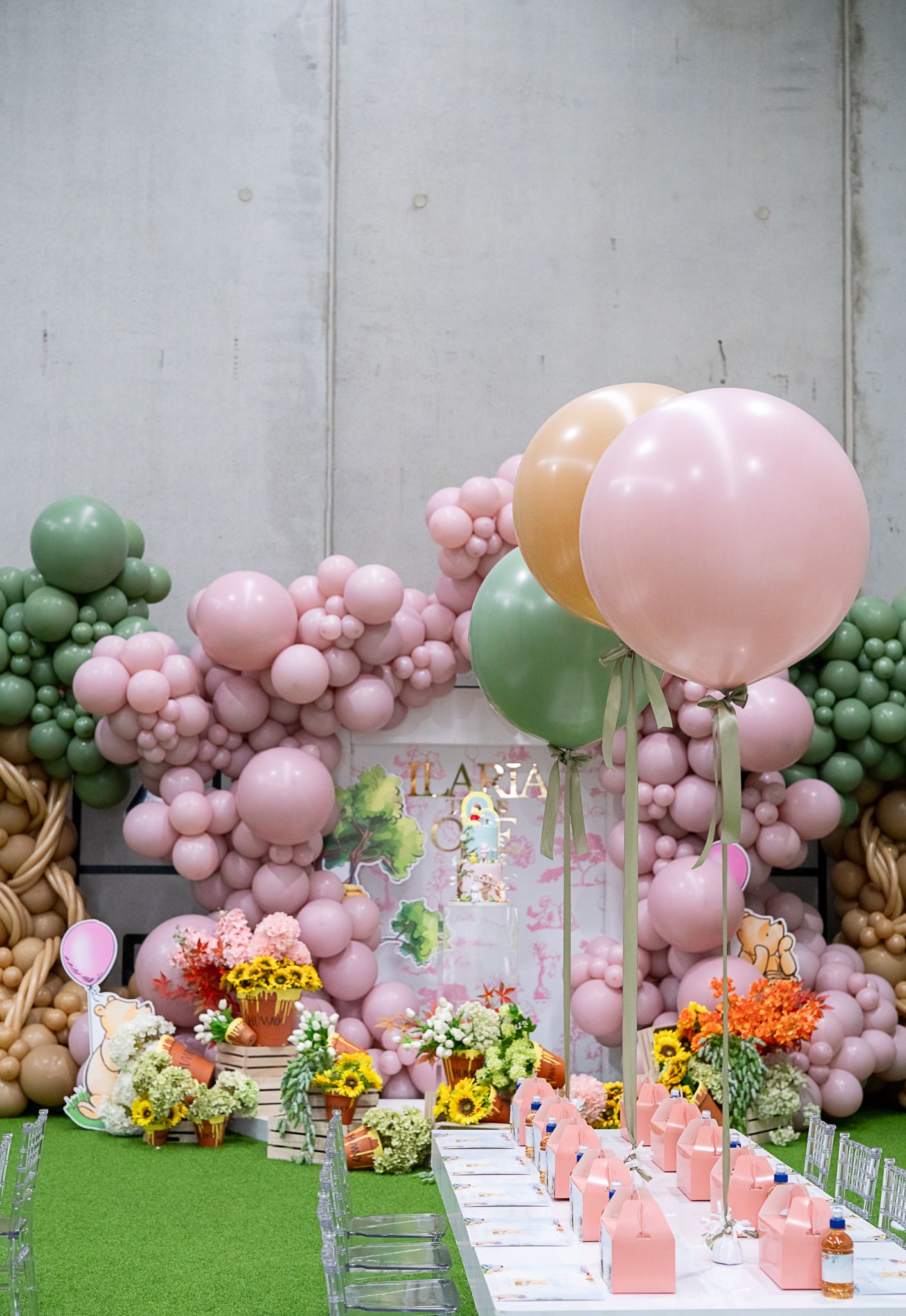 Party setup featuring pink balloon arch, fresh flowers, and table settings on artificial grass.