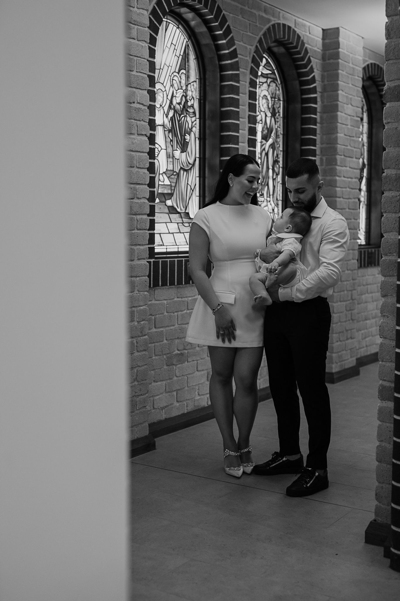 A couple with their baby stands in front of stained glass windows in a church hallway.