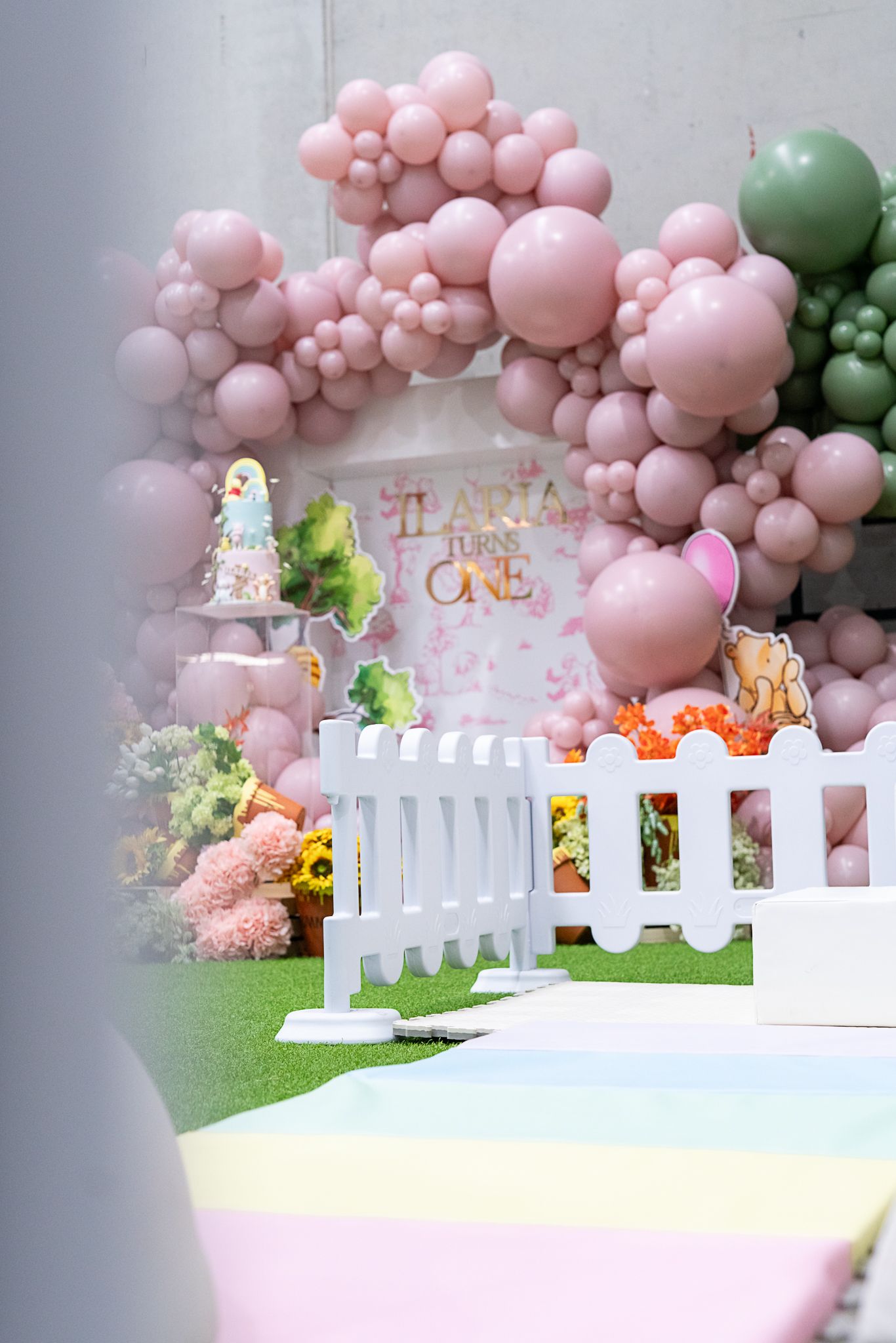 Sweet pink and green balloon arch decorates a whimsical first birthday party setup with white picket fence details.