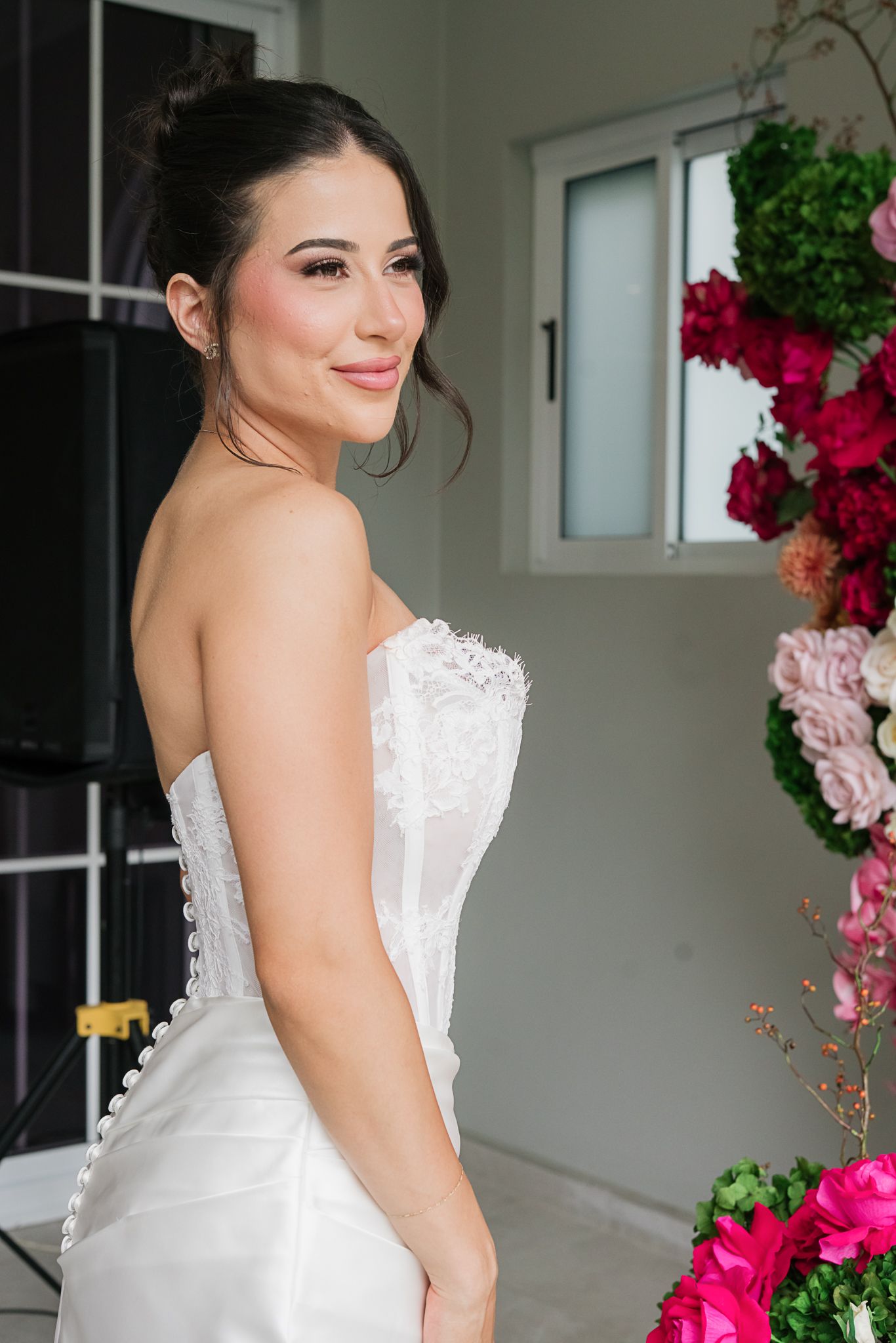 Elegant bride in white strapless lace wedding dress poses next to pink and red floral arrangements.