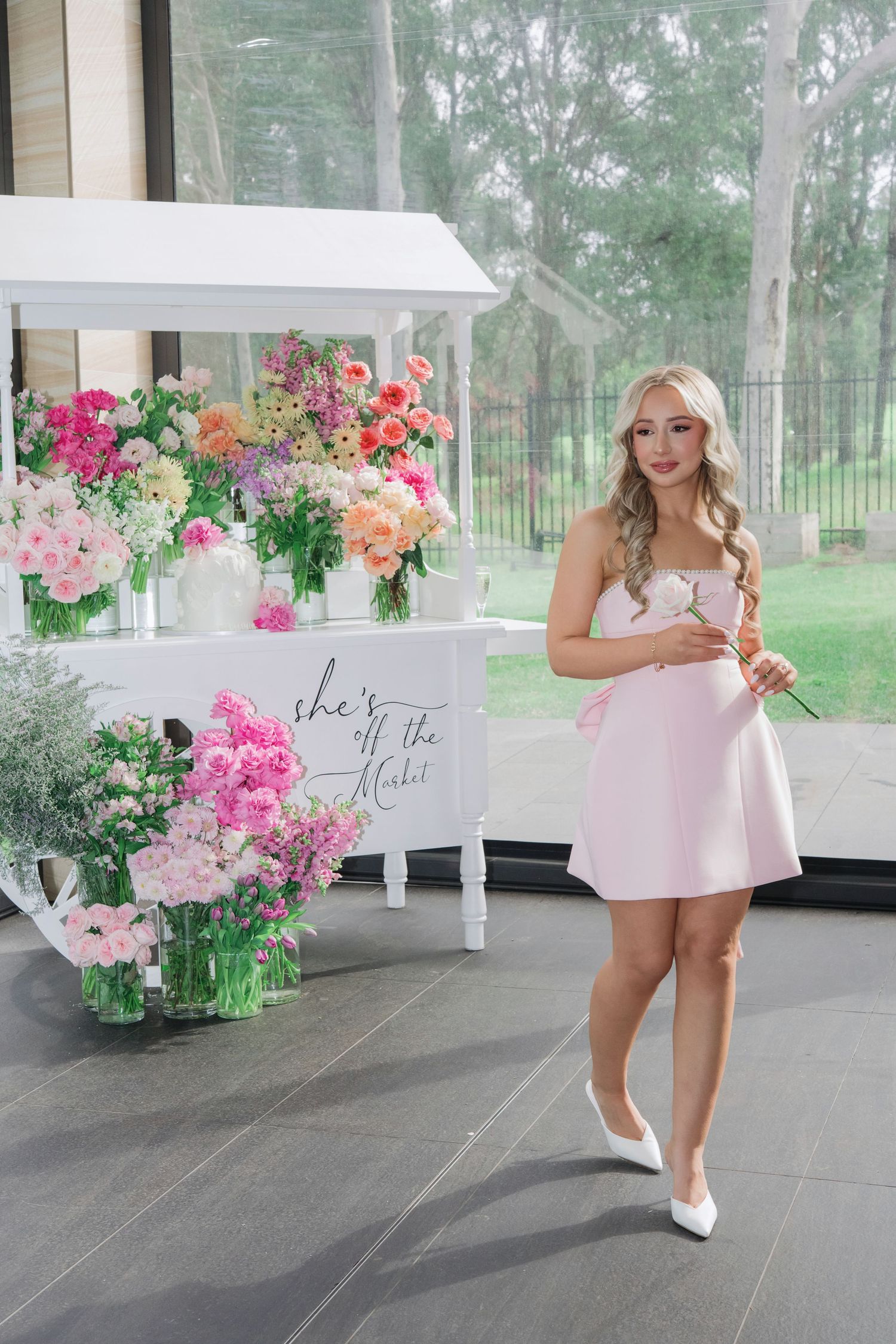 A pink floral display cart with fresh spring arrangements sits beside someone in a light pink dress and white heels.