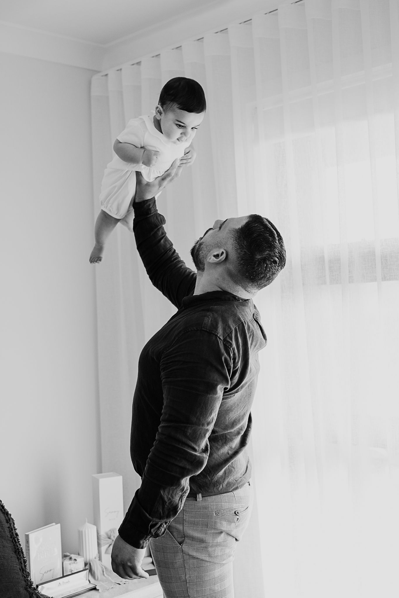 A black and white photo shows a father lifting a small child high in the air while playing together indoors.