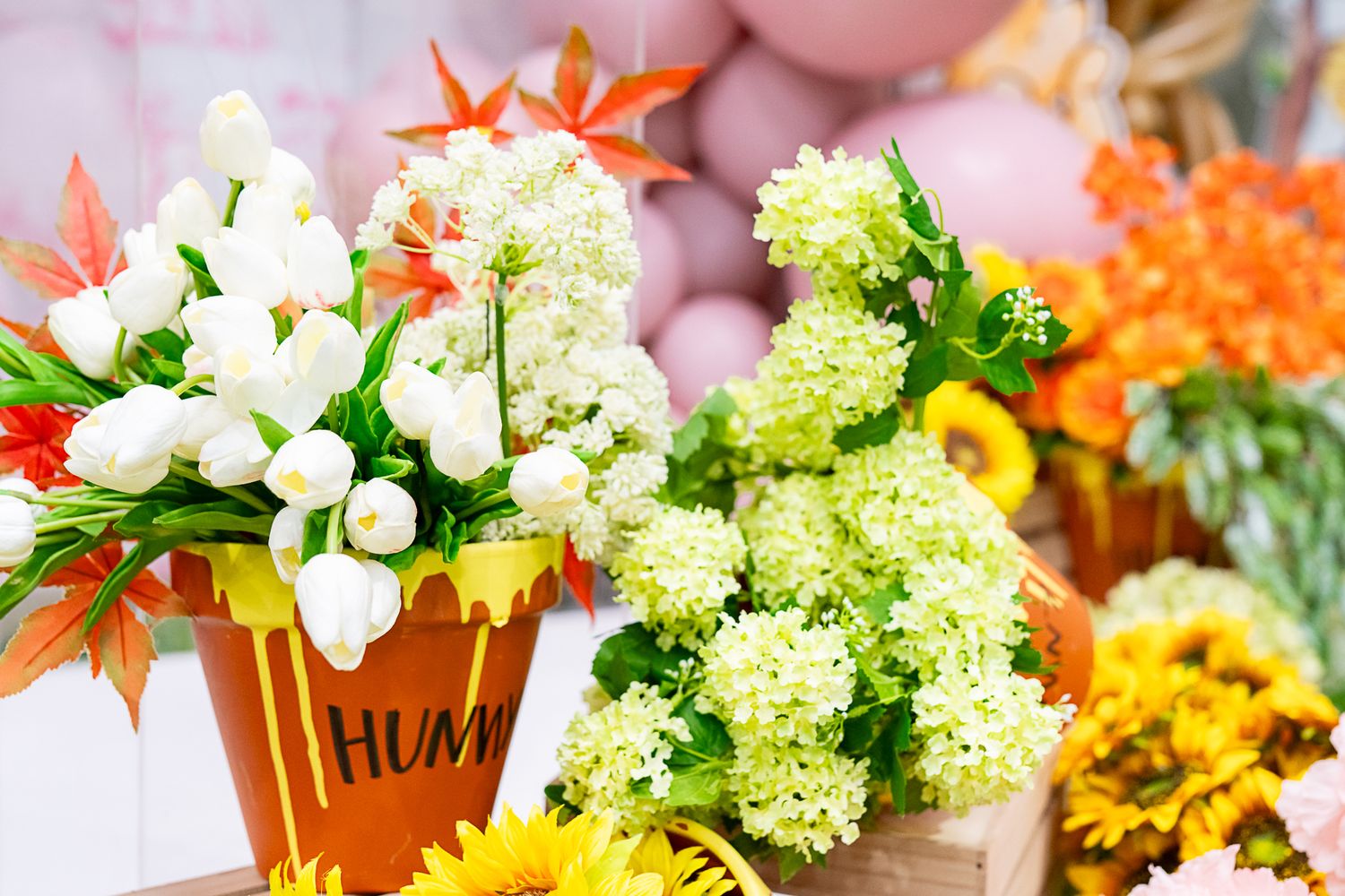 Vibrant floral display with white flowers and greenery in terracotta pots against pink balloon backdrop.