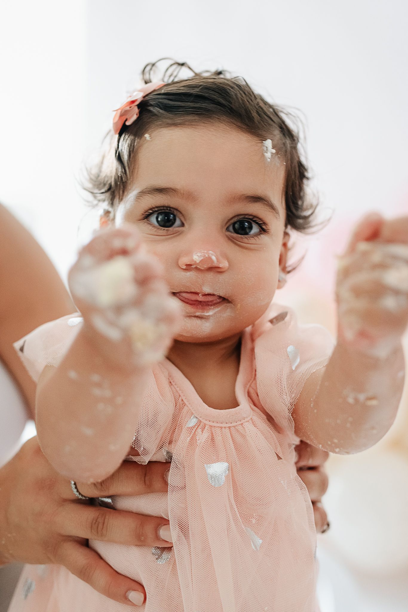 A small child wearing a pink dress reaches out with messy hands during a cake smash photo session.