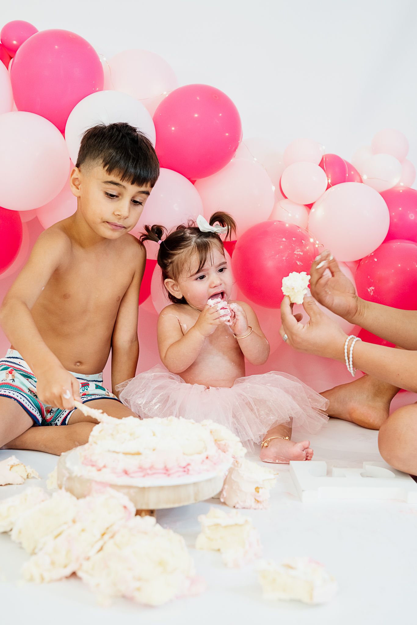 Children enjoying a messy cake smash photo session with pink and white balloons in the background.