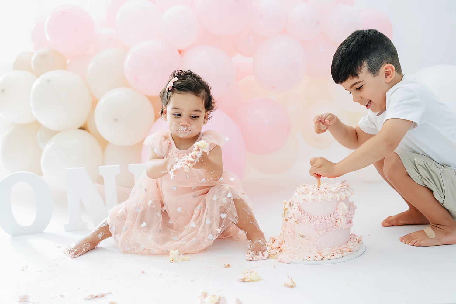 Children enjoying a cake smash photo session against a pink and white balloon backdrop.