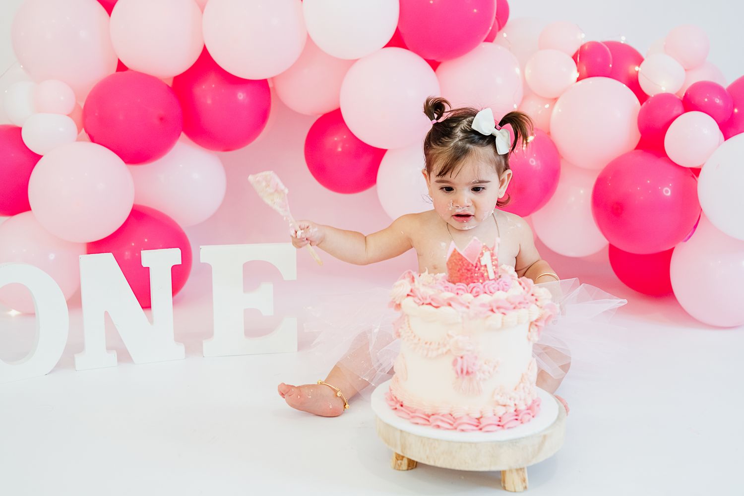 Pink and white balloon backdrop with ONE letter decor and small child in tutu dress celebrating with birthday cake.
