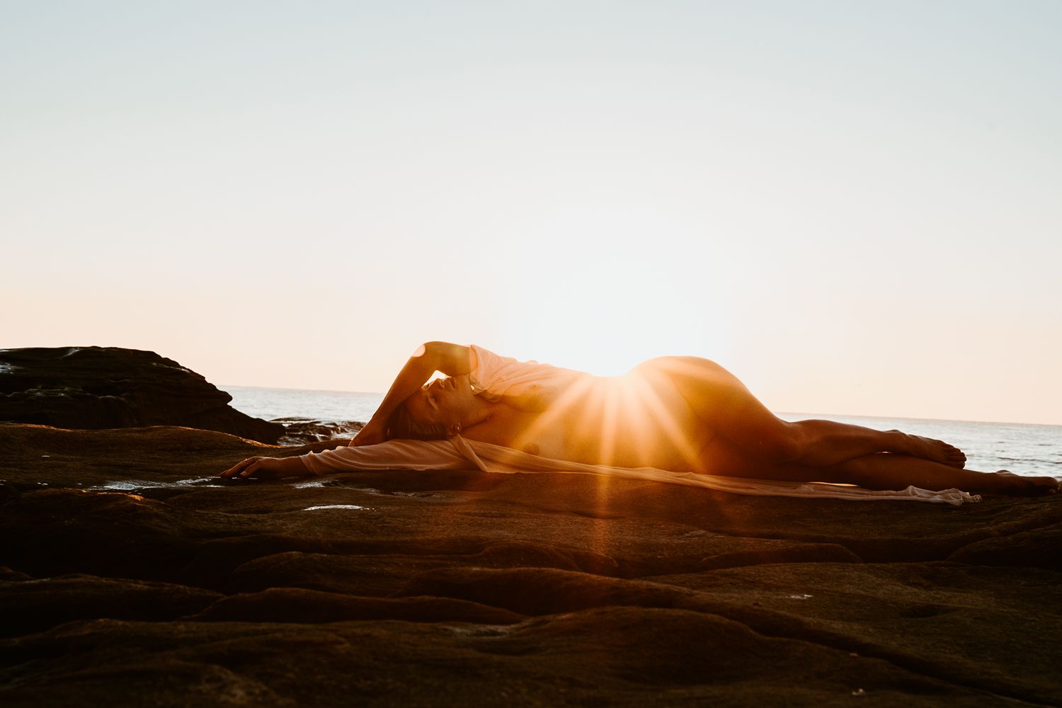 Person posing on coastal rocks during dramatic sunset.