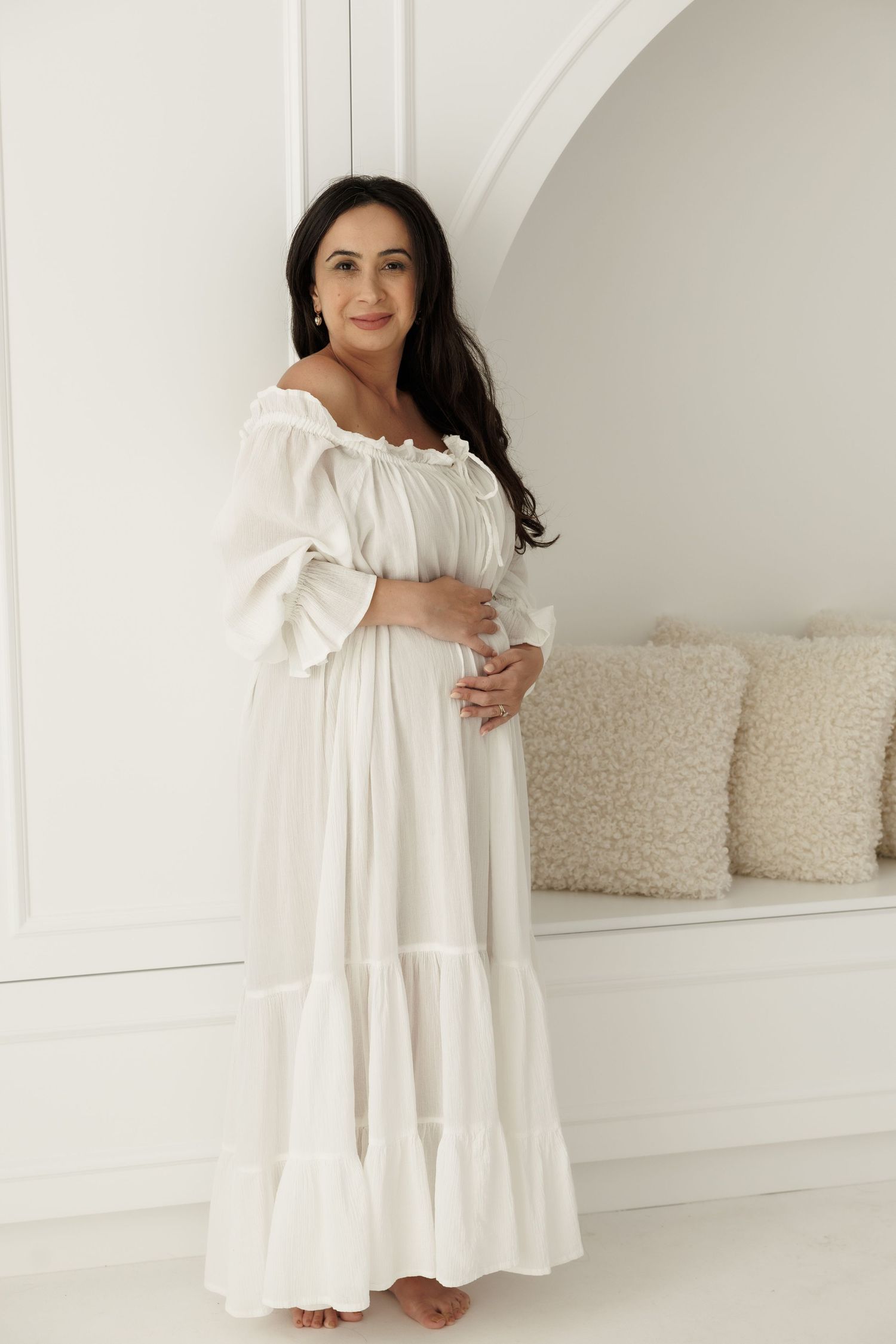 A pregnant woman in a flowing white off-shoulder maxi dress poses against a white wall with decorative pillows.