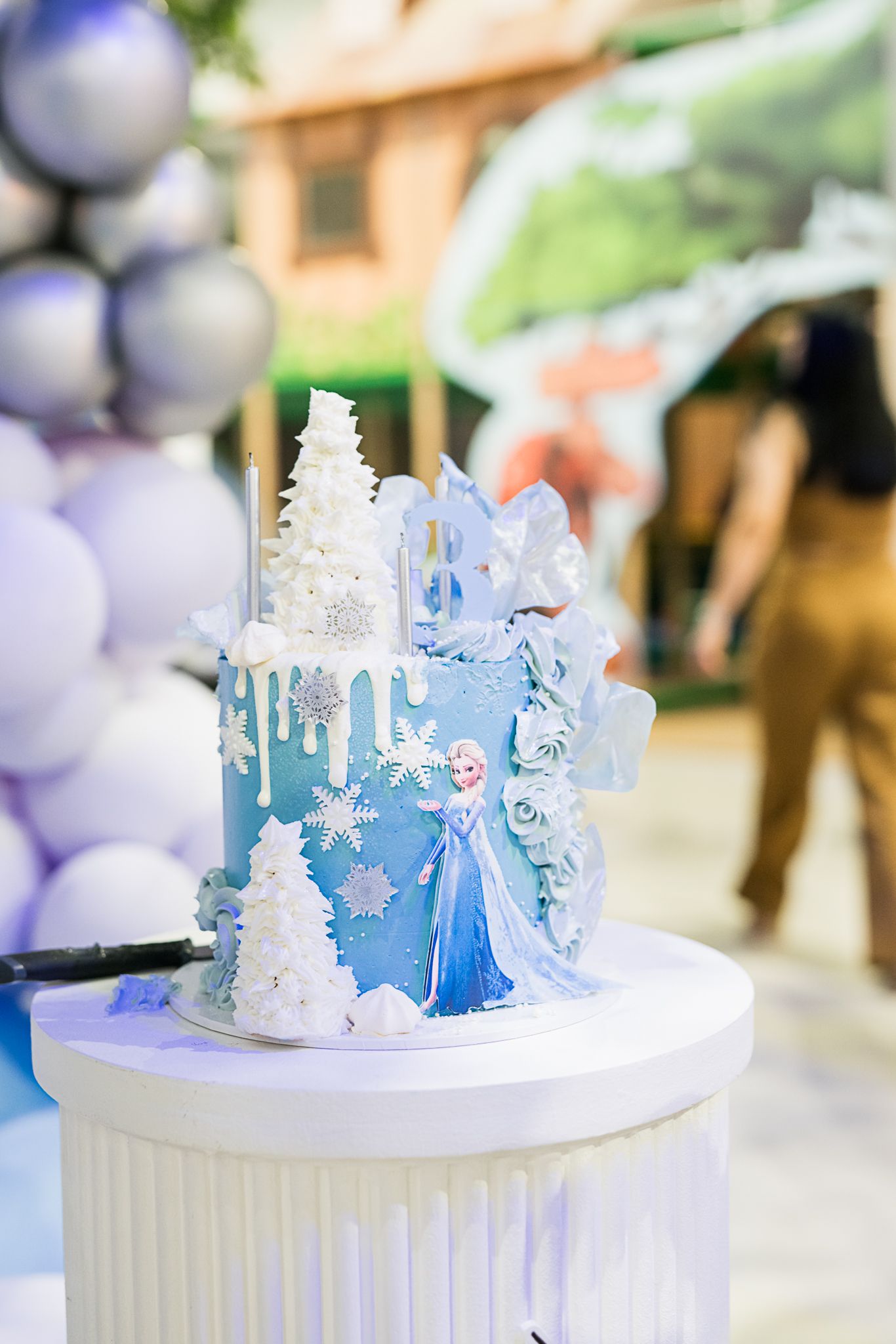 Frozen-themed birthday cake decorated with white icing, blue drips, and snowflake designs on a white pedestal.