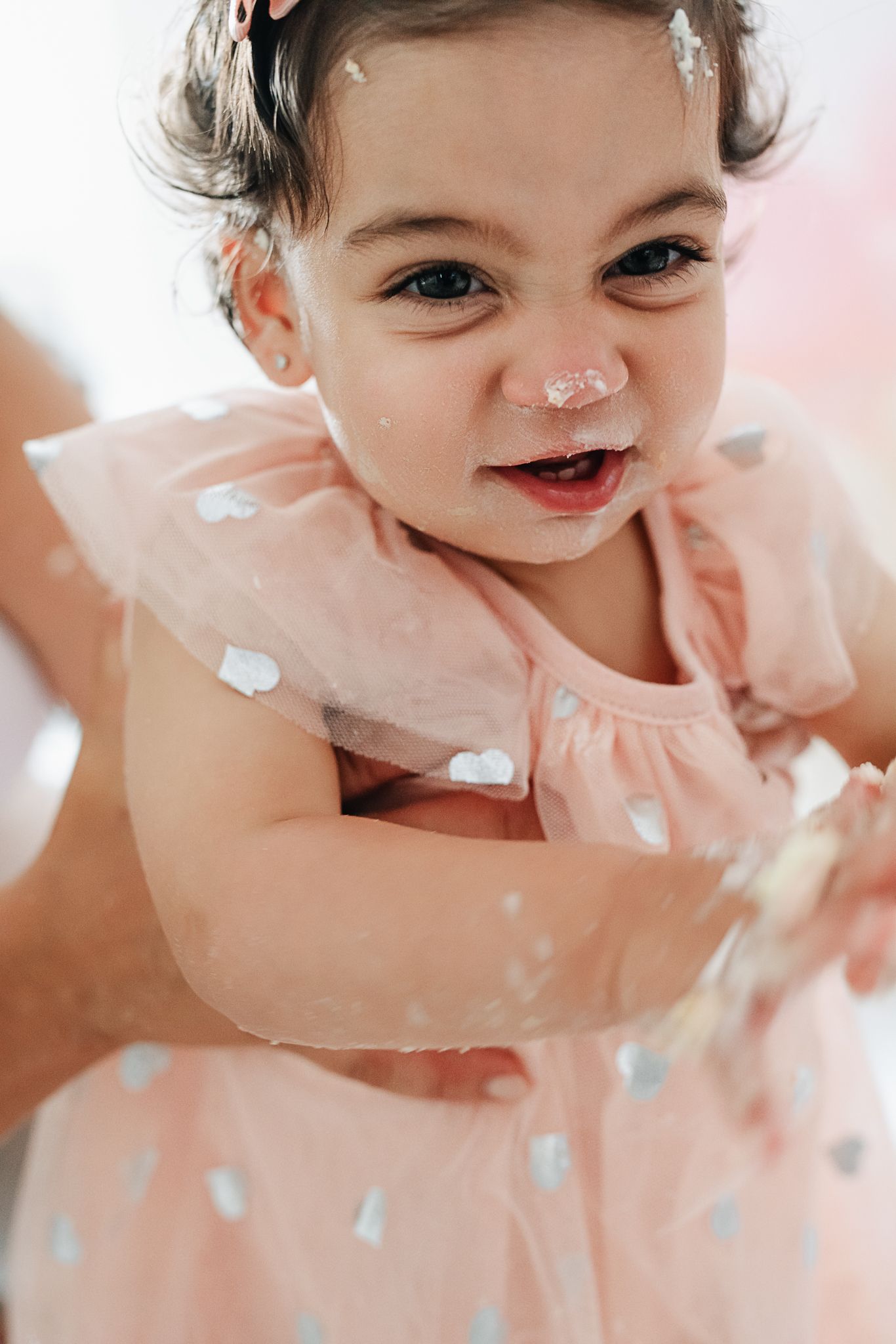 Joyful toddler in pink polka dot dress laughs while covered in cake.