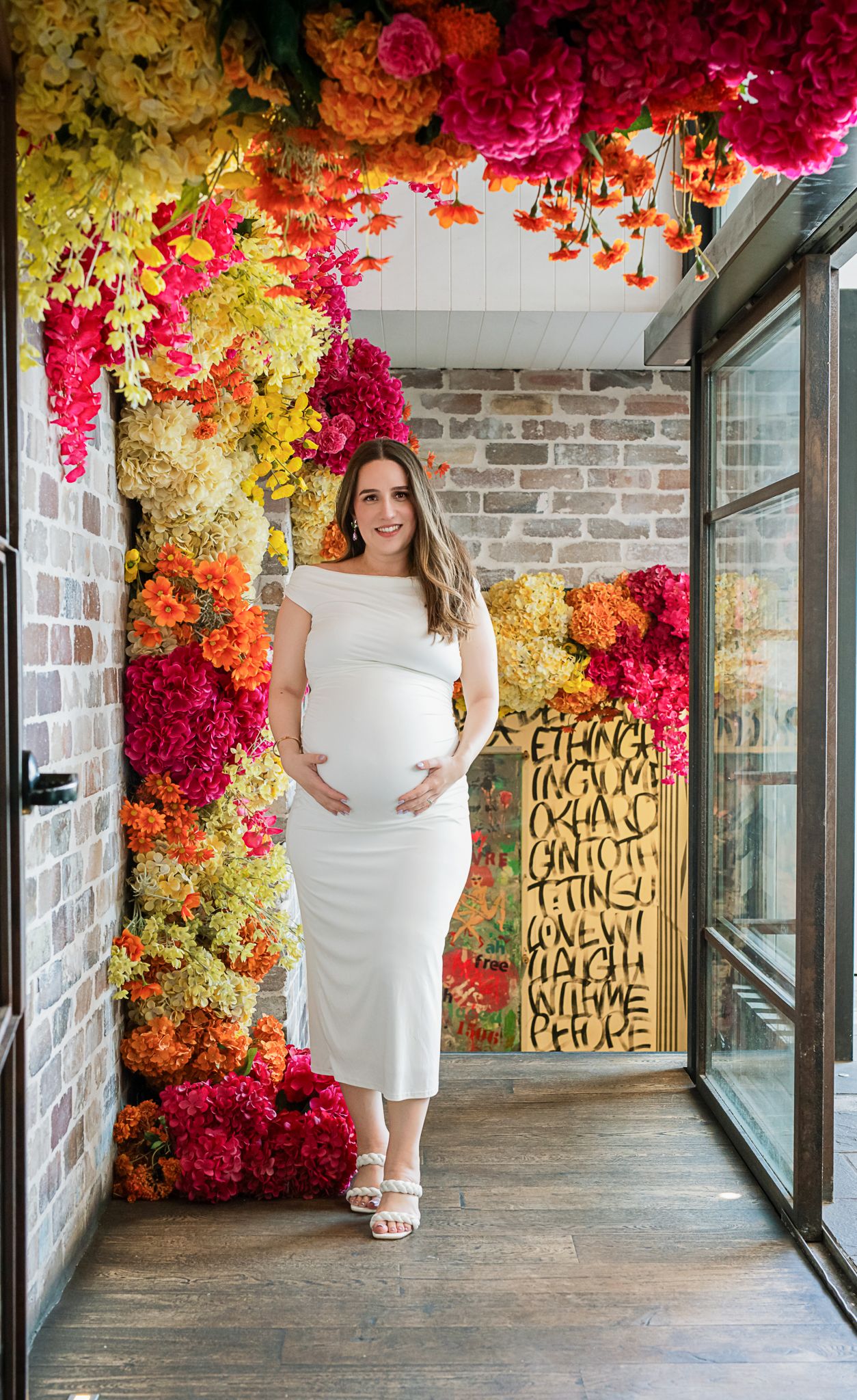 A maternity photo shoot featuring a white dress against a vibrant floral backdrop of red, orange and yellow blooms.