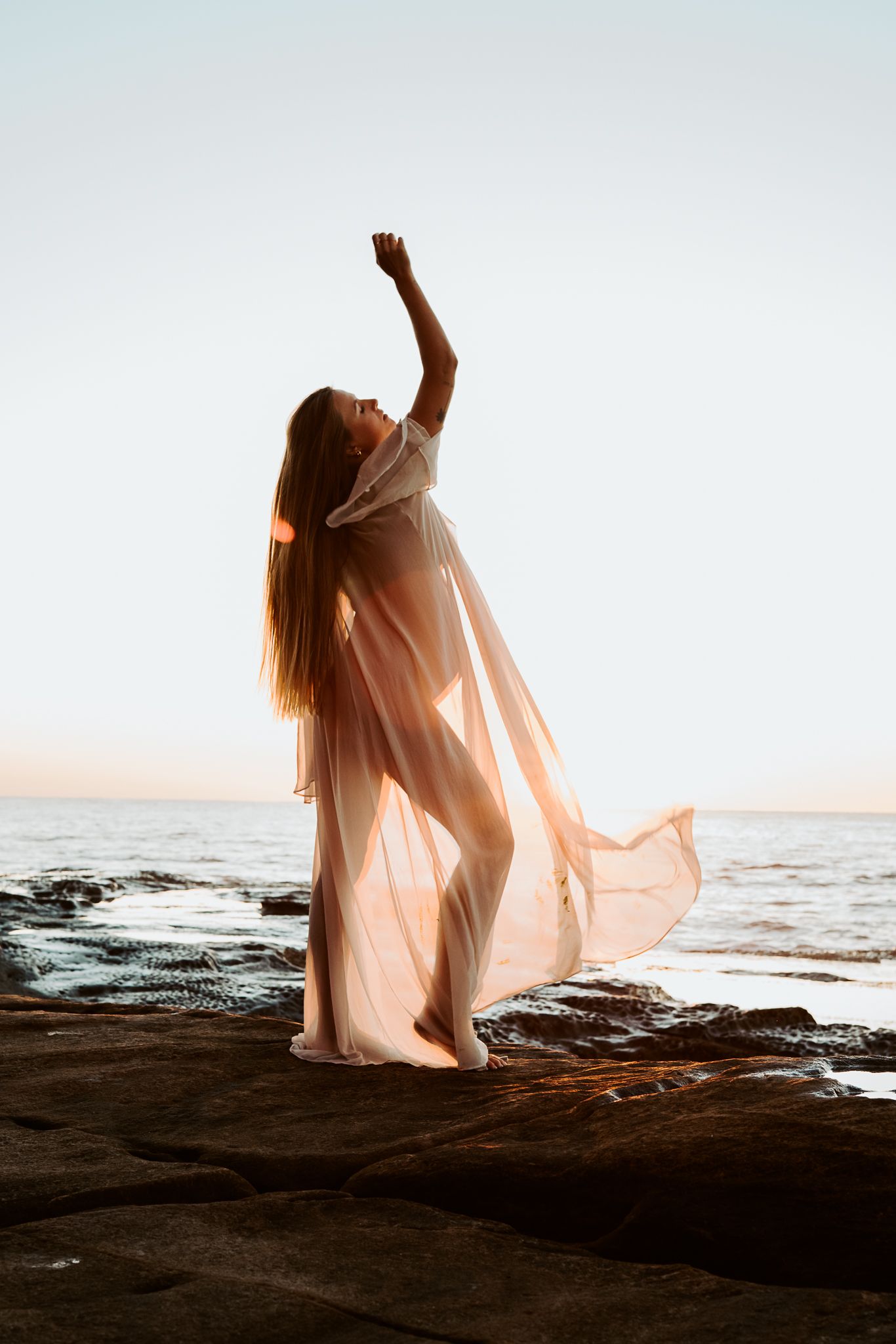 Figure in flowing white dress reaches skyward during coastal sunrise maternity photoshoot.