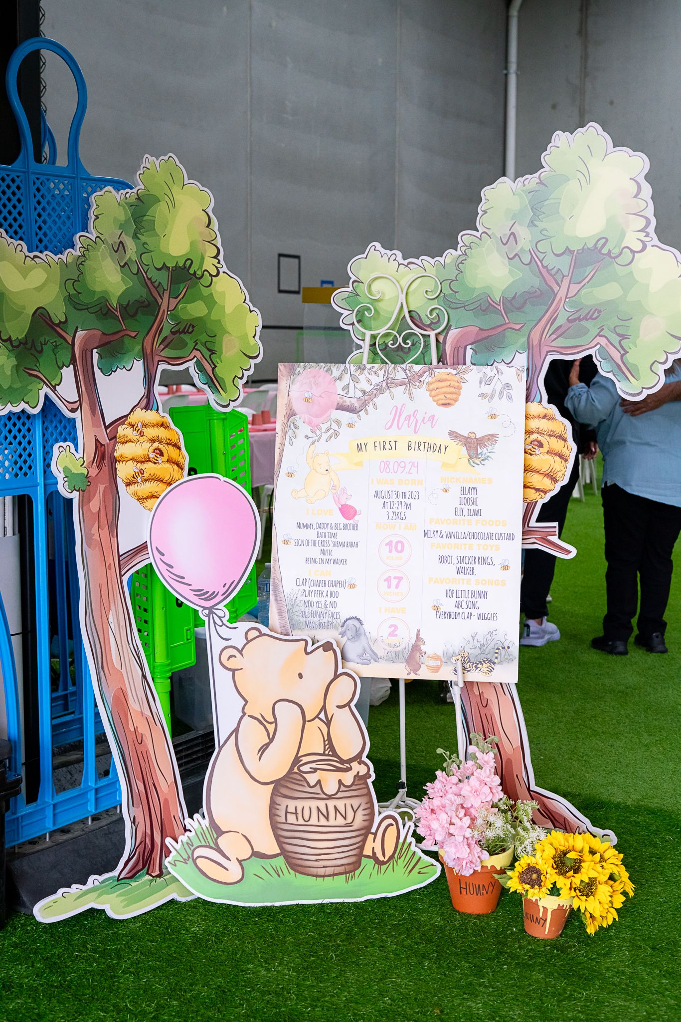 Whimsical party setup with cartoon bear cutout, trees, and decorative signage on artificial grass.