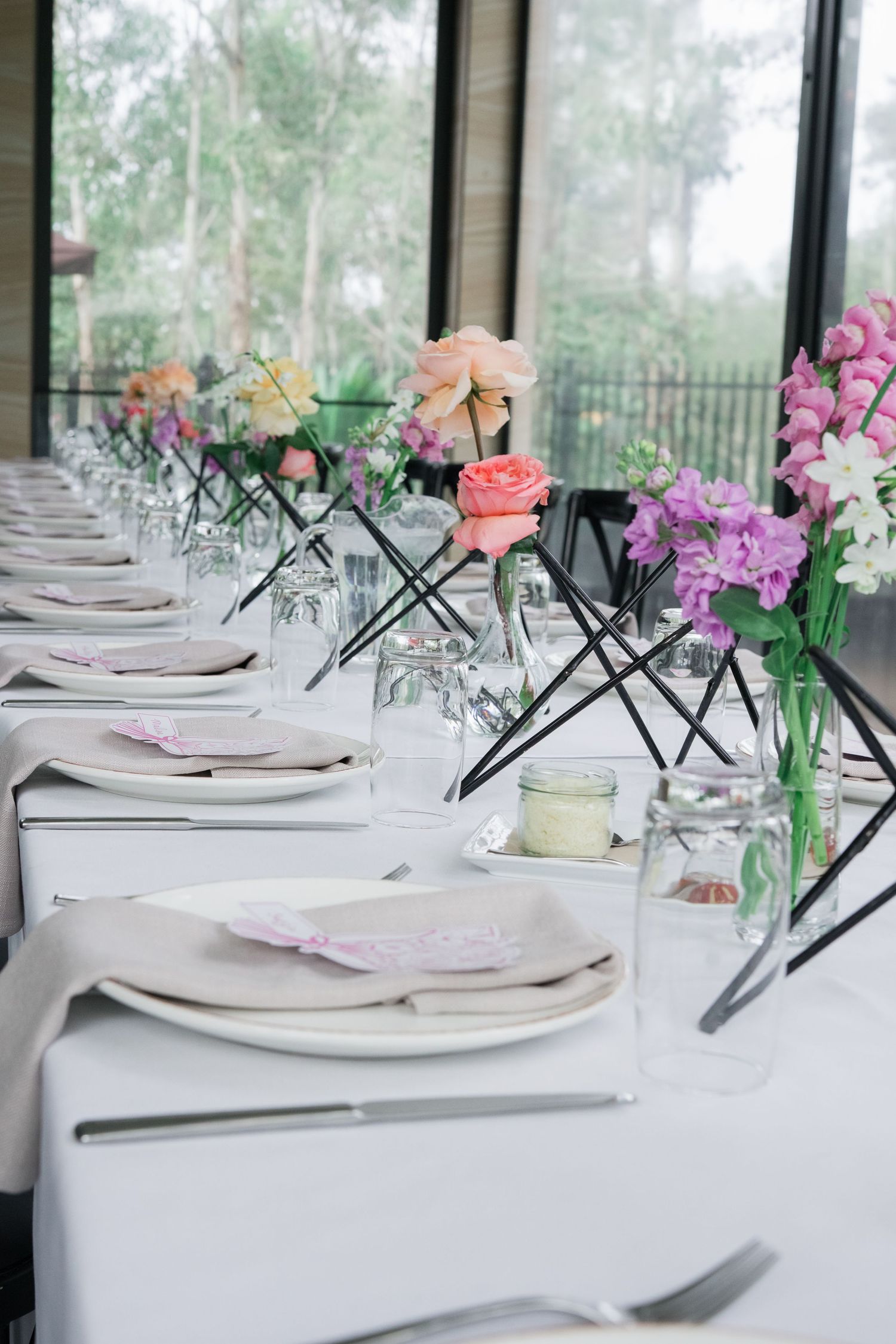 Elegant table setting with white linens and delicate floral centerpieces featuring pink, purple and peach blooms in glass vases.