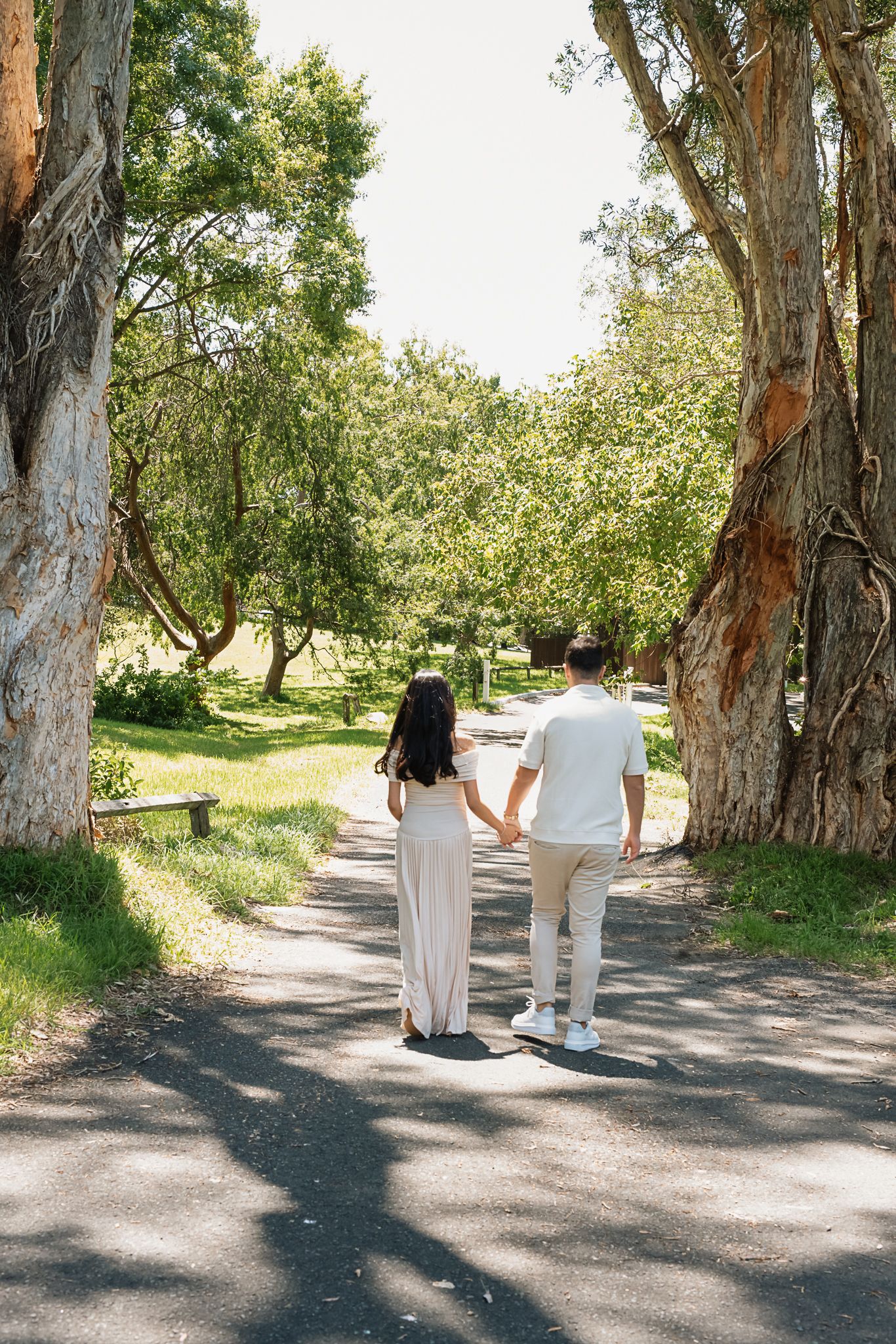 Couple holding hands walking down tree-lined path in matching cream outfits during maternity shoot.